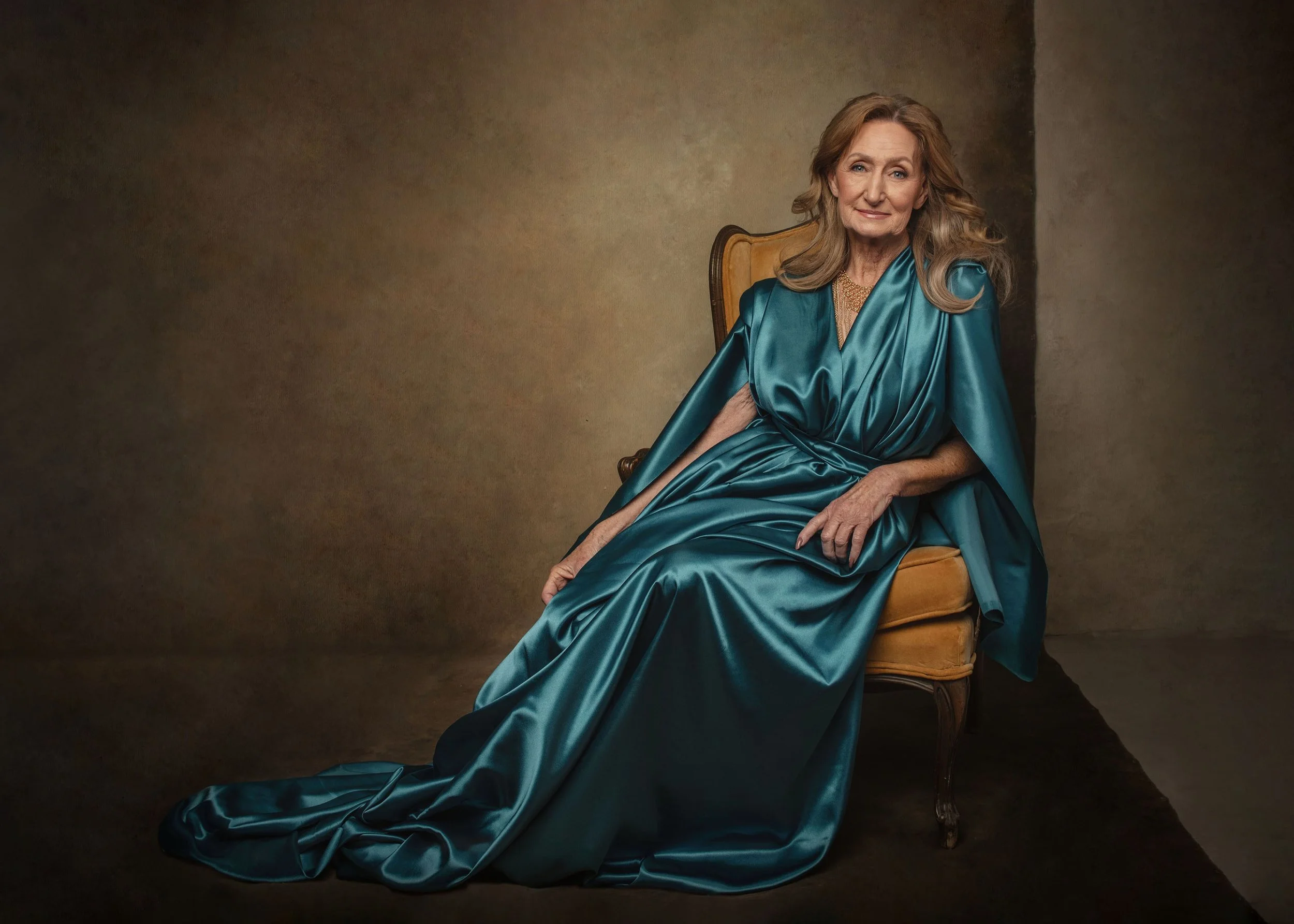 An elderly woman with long gray hair, wearing a shiny teal dress, sitting on a vintage orange and wood chair.