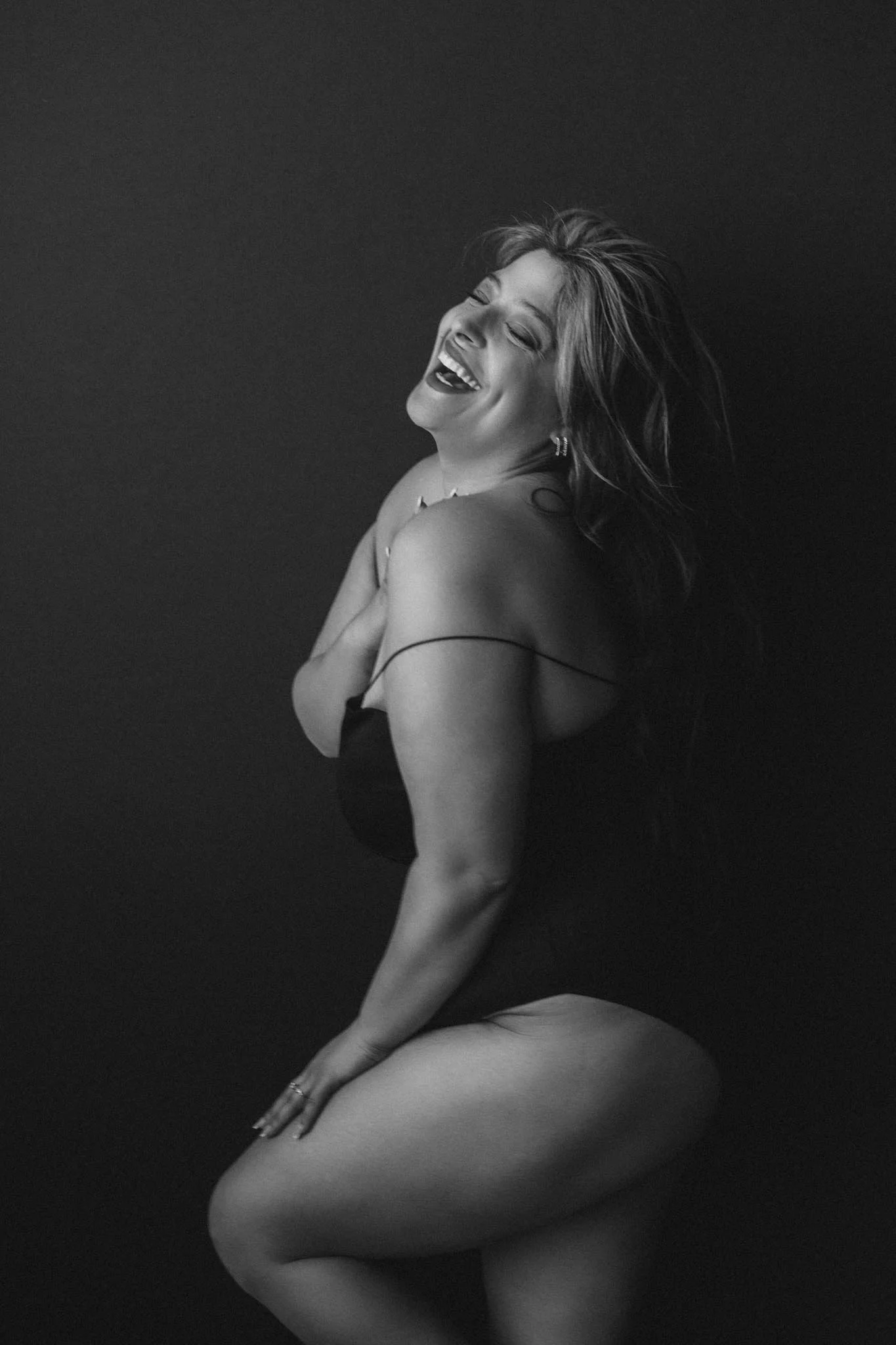 Black and white portrait of a woman laughing, posing against a dark background. She is wearing a spaghetti strap top and appears to be seated, with her arms crossed over her chest and her head tilted back.
