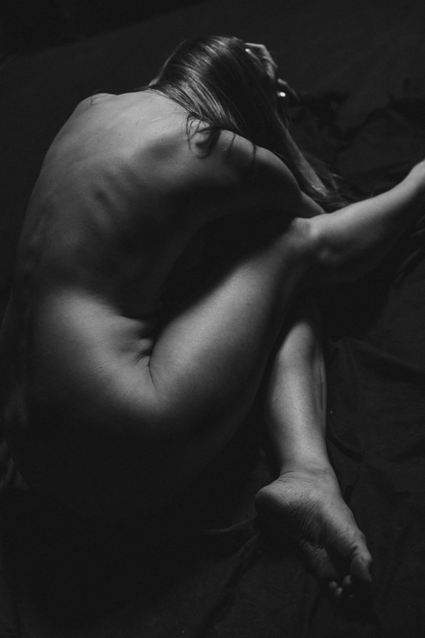 A black and white photograph of a nude woman sitting on a dark surface with her knees pulled up to her chest, her arms wrapped around her legs, and her head resting on her knees. Her face is not visible, and her long hair covers her face.