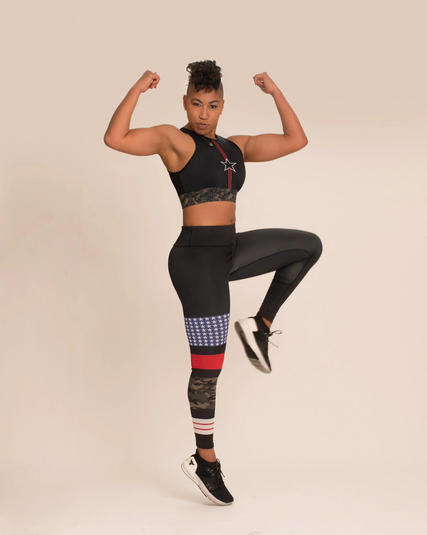A woman flexing her biceps and standing on one leg, wearing black athletic clothing and sneakers, against a plain beige background during her editorial portrait session.