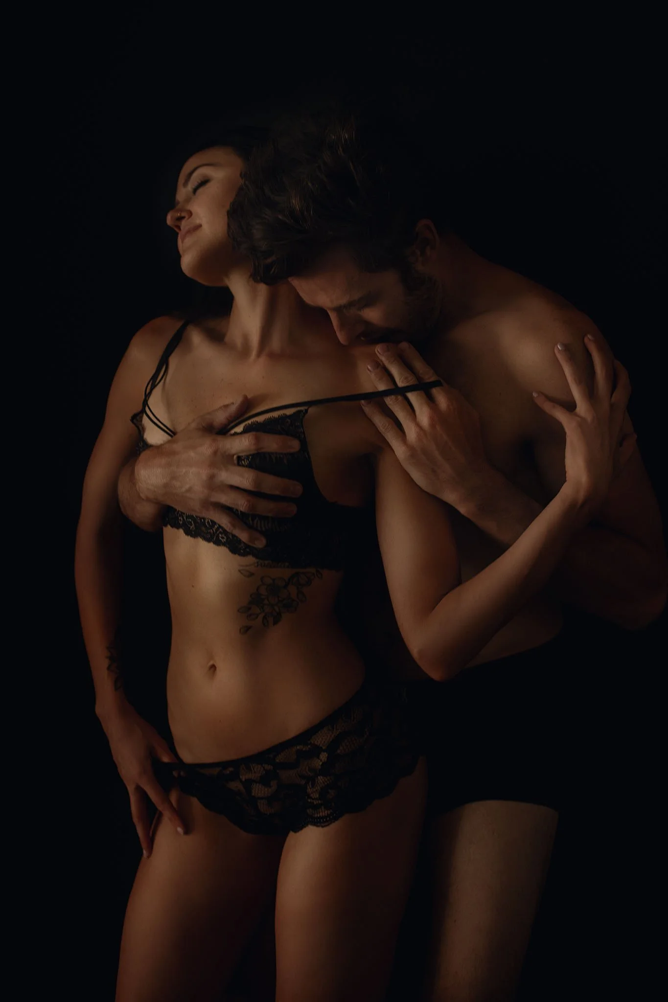 A woman and man embrace in lingerie and are set against a dark background.