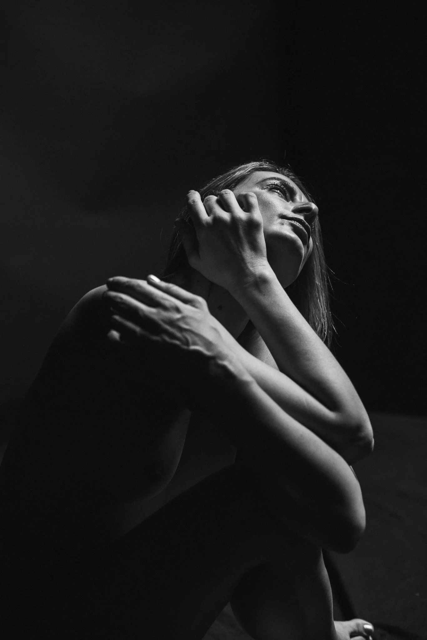Black and white photo of a woman sitting with her knees drawn up, her head tilted back, touching her face with her hand, and looking upward, against a dark background.
