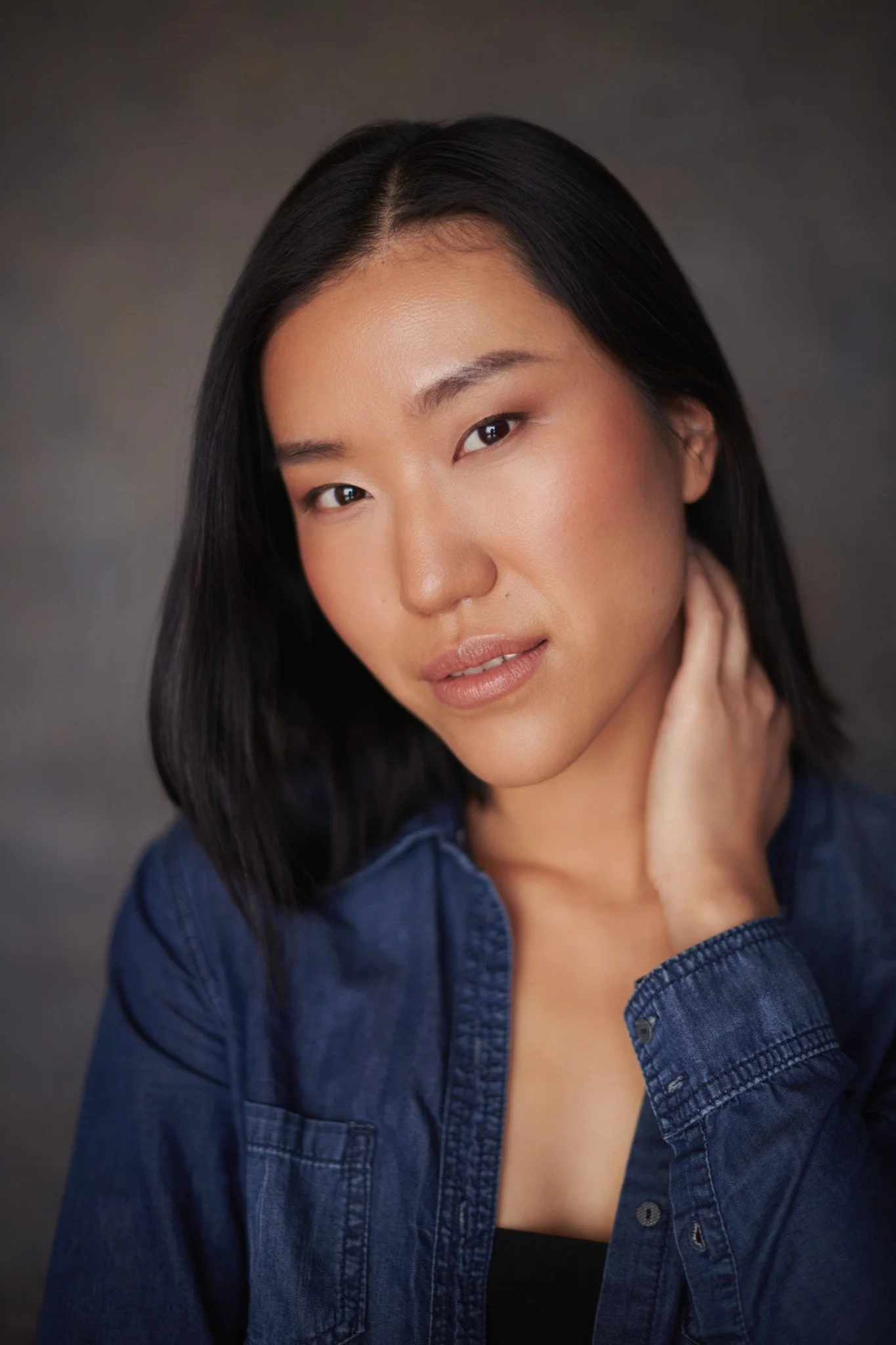 A portrait of an Asian woman with black hair, wearing a denim shirt, looking confidently at the camera with her hand touching her neck.