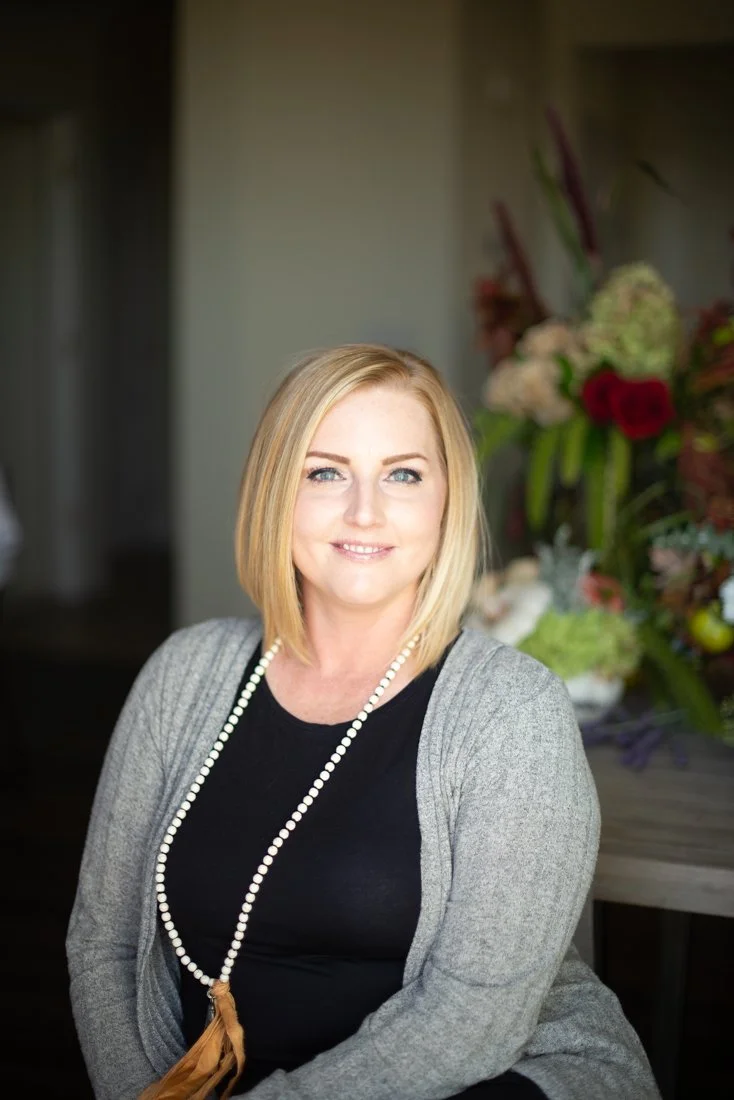 A woman with shoulder-length blonde hair and blue eyes smiling at the camera, wearing a black top, a grey cardigan, and a pearl necklace, with a background of a large floral arrangement.