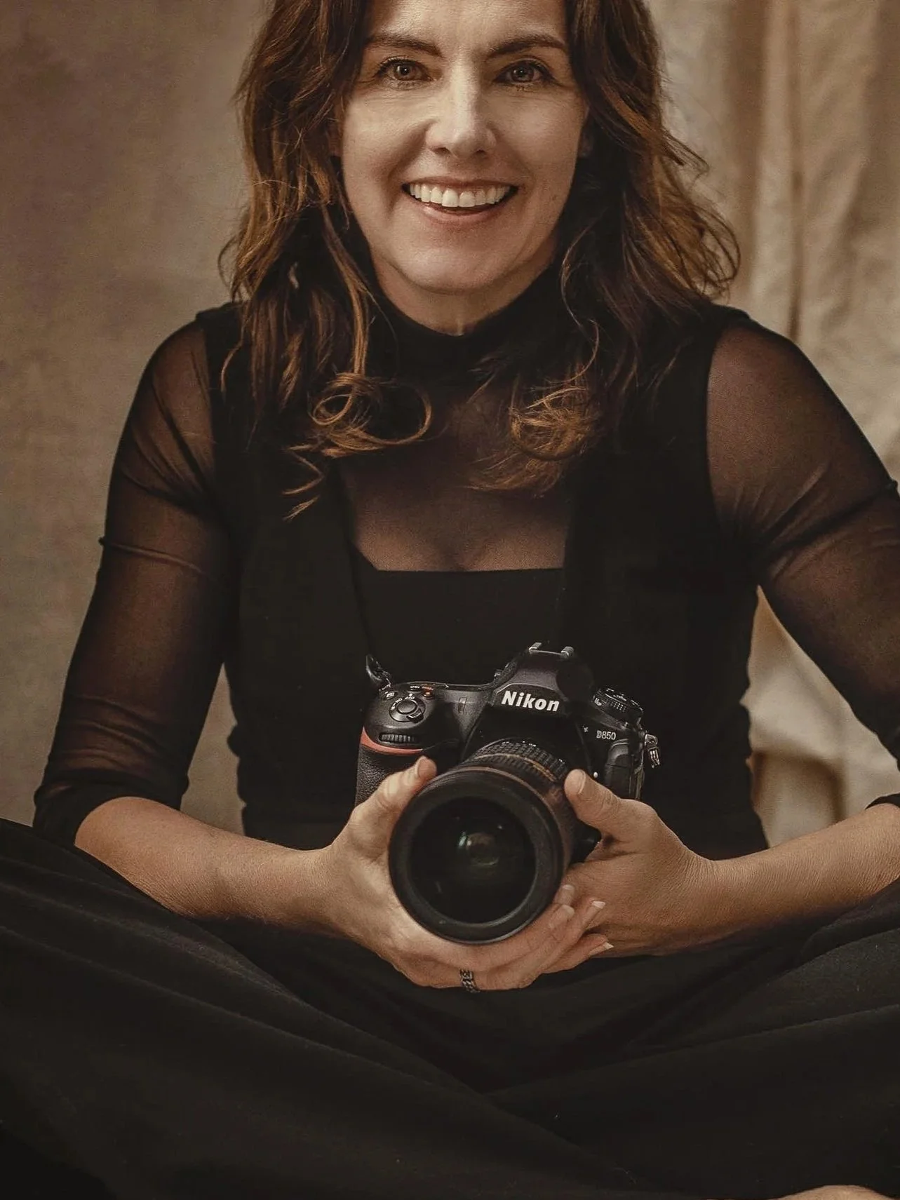 Woman with brown hair holding a Nikon camera, smiling at the camera, wearing a black top with sheer sleeves.