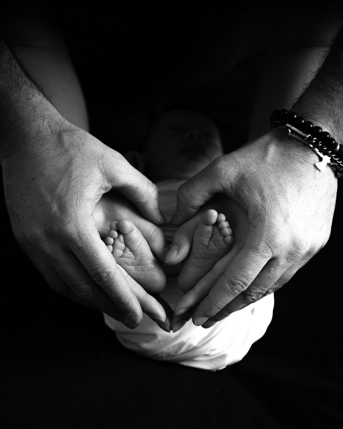 Black and white photo of two adult hands forming a heart shape around a baby's feet, with a baby lying beneath, blurred face in the background.