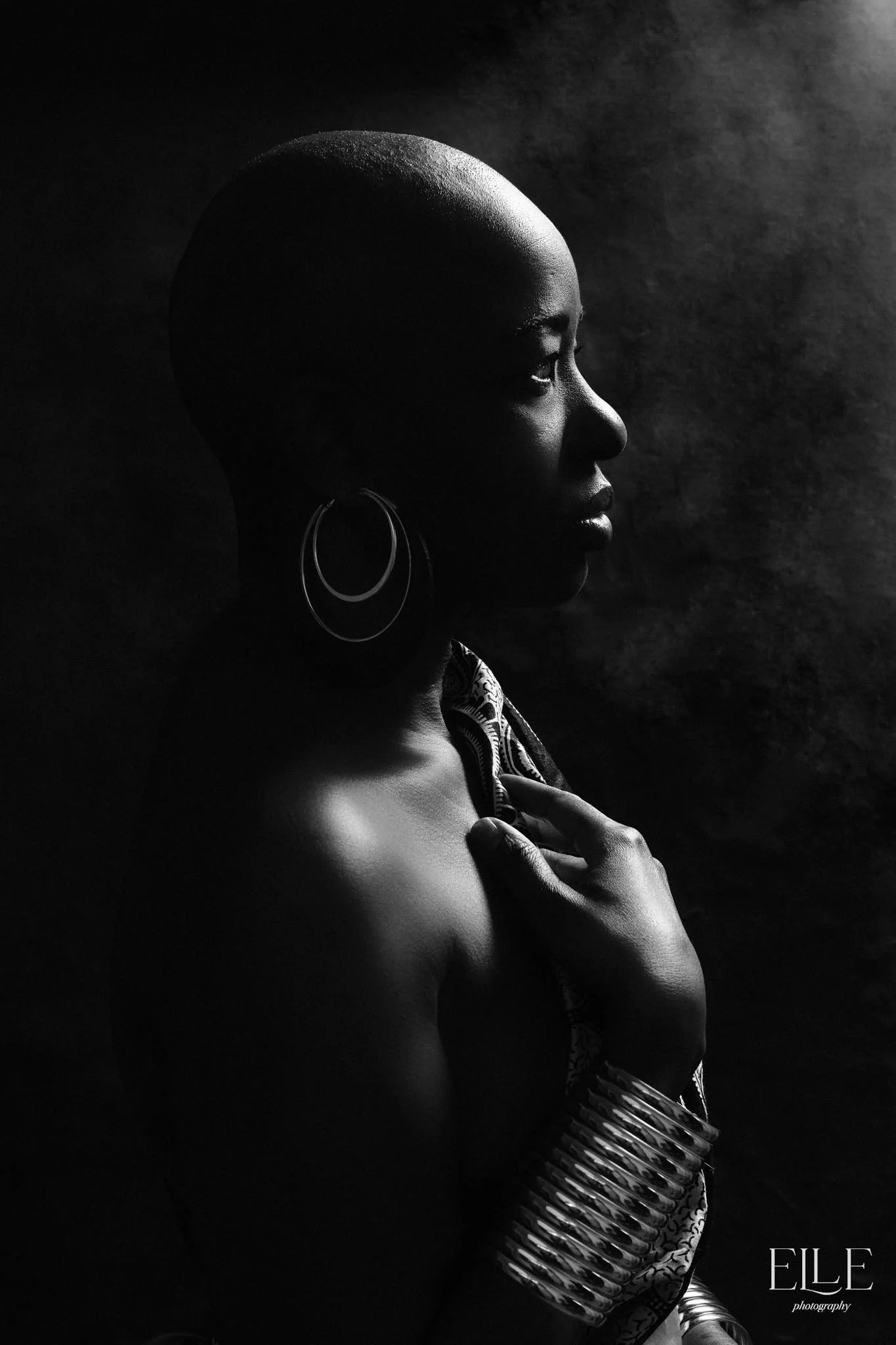 Black and white portrait of a woman with a shaved head, wearing hoop earrings and a patterned scarf, with one hand on her chest and jewelry on her wrist, looking to the side.