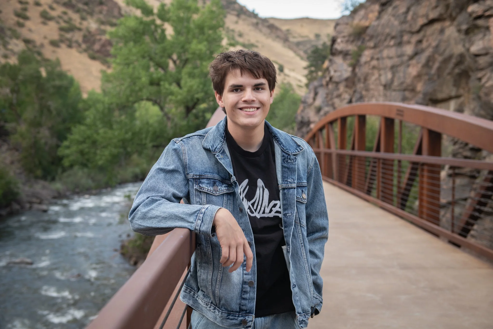 A young man with short brown hair smiling while leaning on a bridge railing. He is wearing a denim jacket over a graphic T-shirt. The background features a flowing stream, green trees, and rocky hills in a natural outdoor setting.