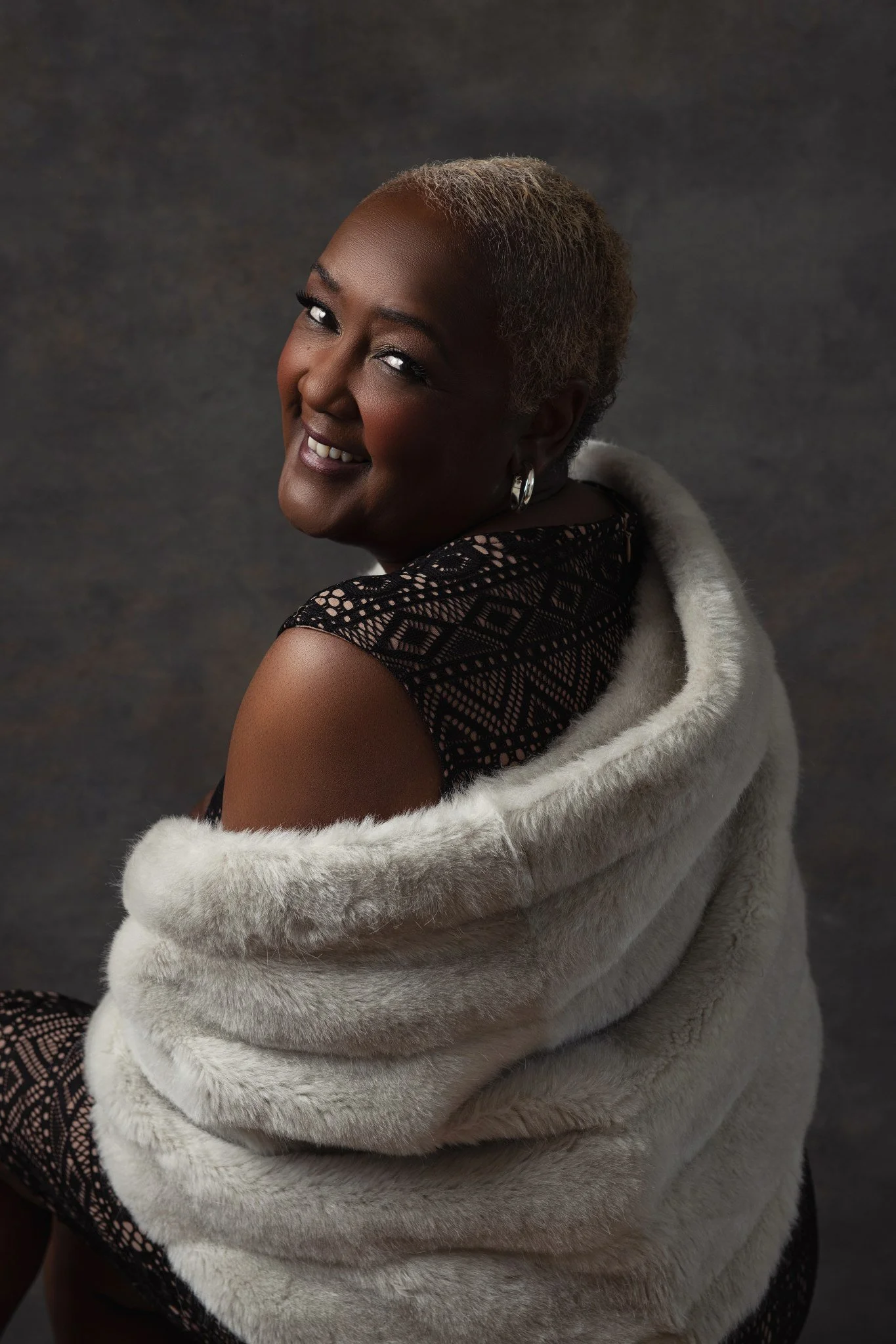Portrait of a smiling middle-aged woman with short gray hair, wearing a black lace dress and silver earrings, wrapped in a white fur coat against a dark background.