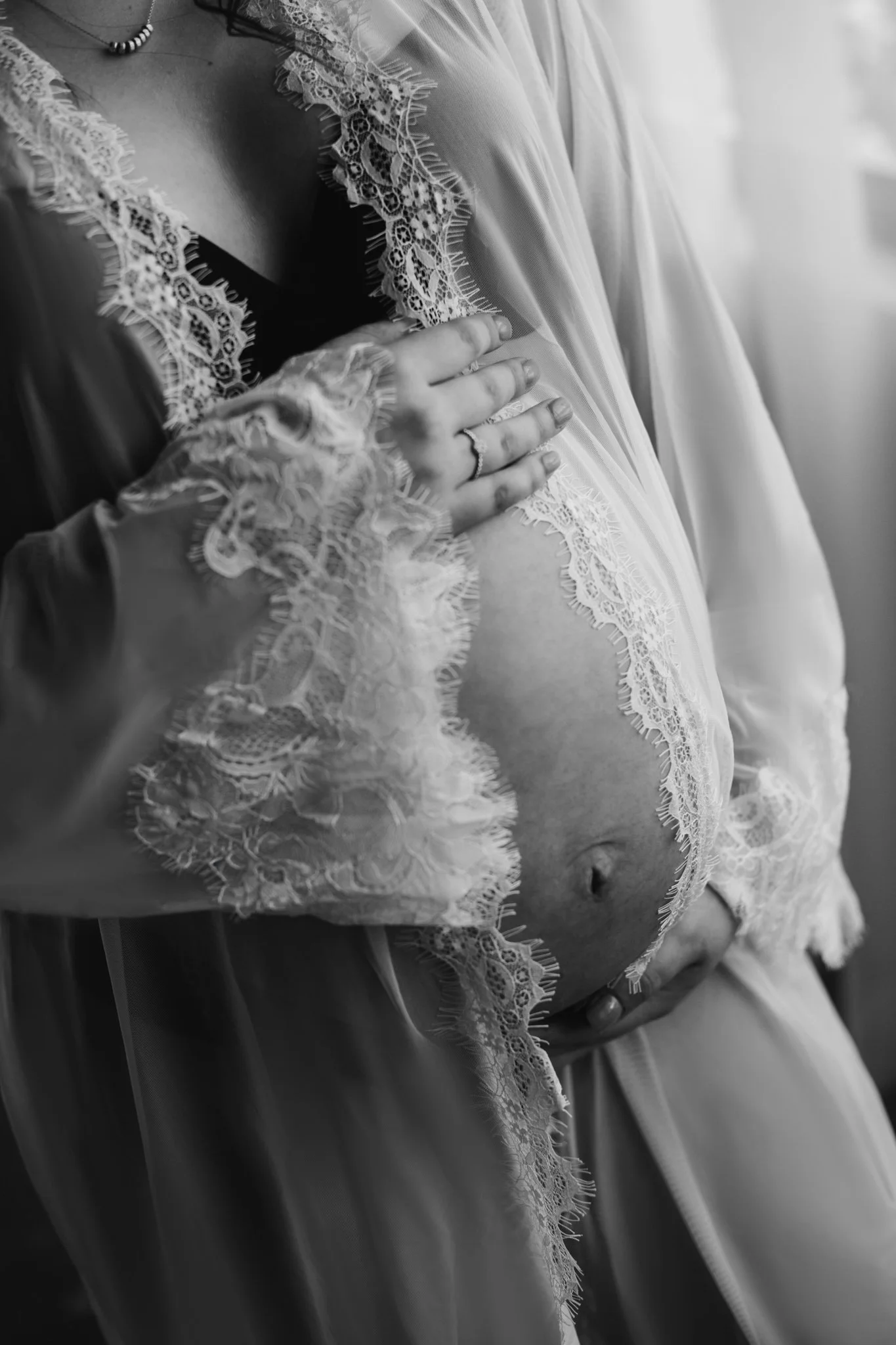 Close-up of a pregnant woman wearing lace and satin robes, with one hand resting on her belly and a ring on her finger, in black and white.