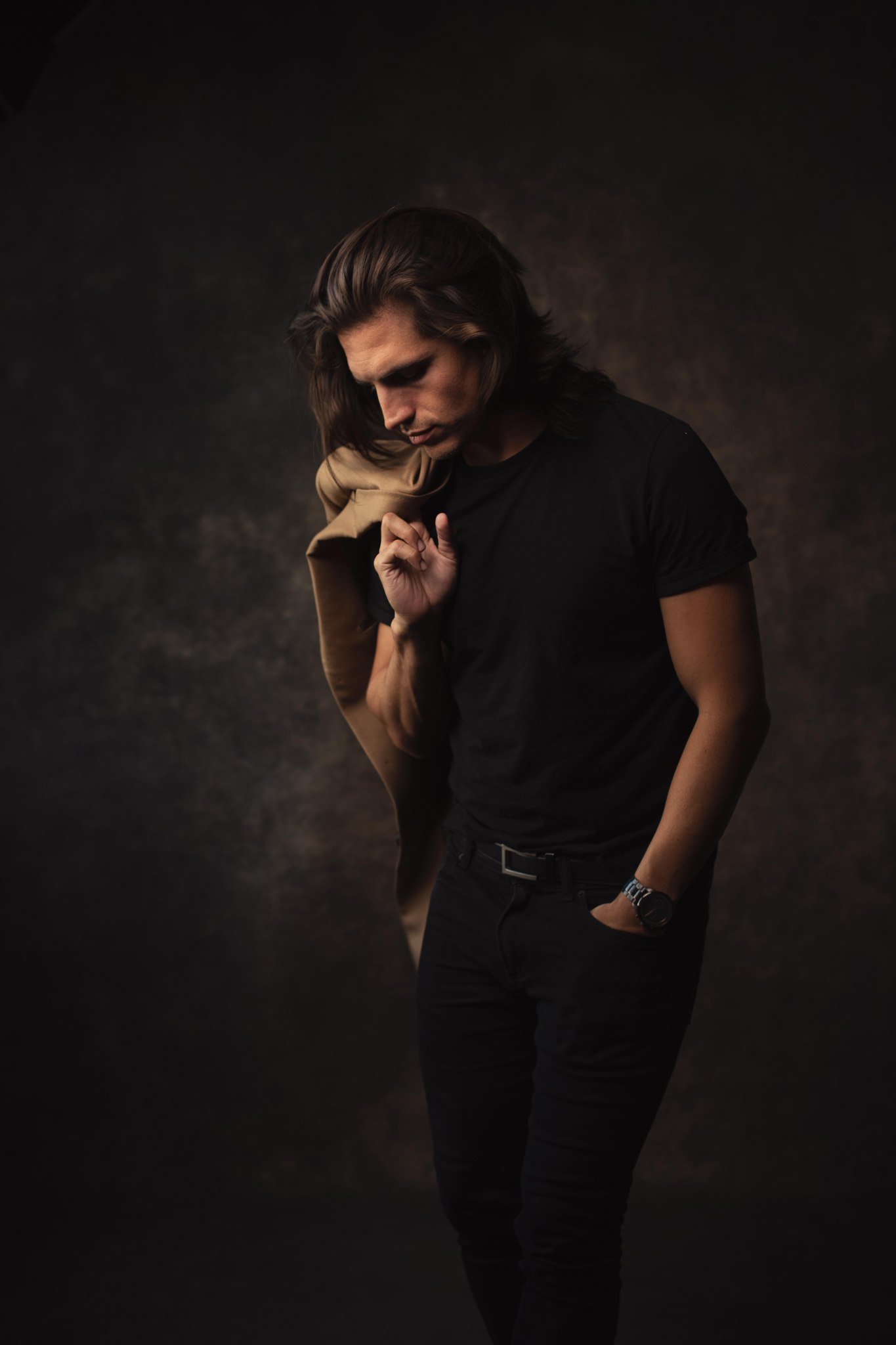 A young man with long hair wearing a black t-shirt, dark pants, and a wristwatch, holding a tan jacket over his shoulder, looking down thoughtfully against a dark textured background.