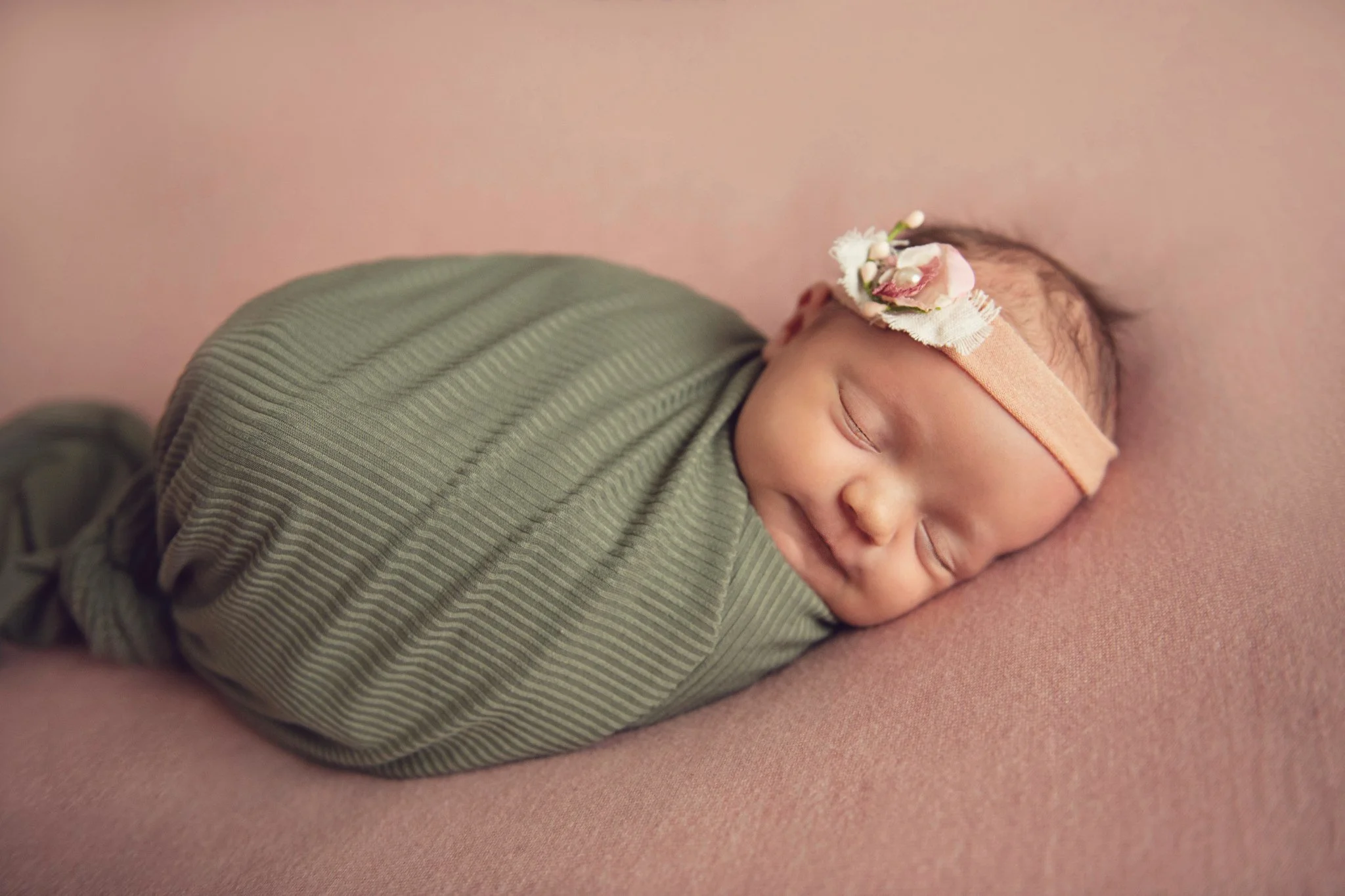 A sleeping baby wrapped in a green fabric on a pink surface, wearing a peach headband with a flower embellishment.