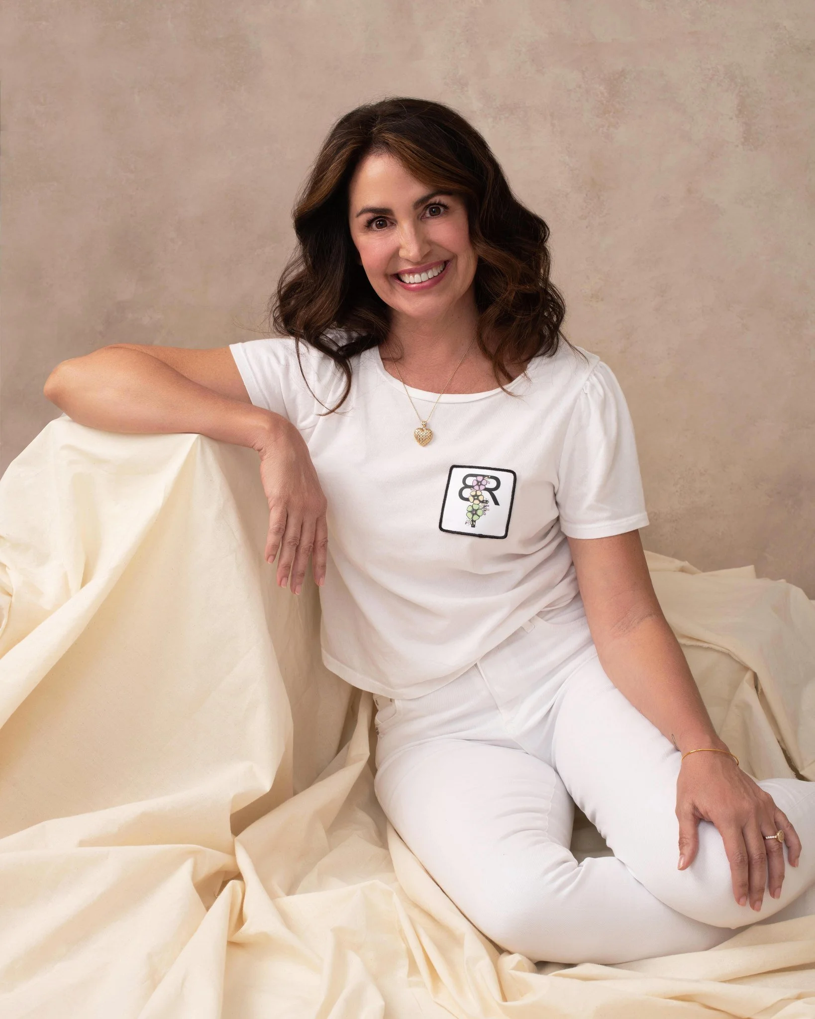 A woman with dark wavy hair sitting on cream-colored fabric, wearing a white t-shirt with a patch featuring a letter and a flower design, and white pants, smiling at the camera.