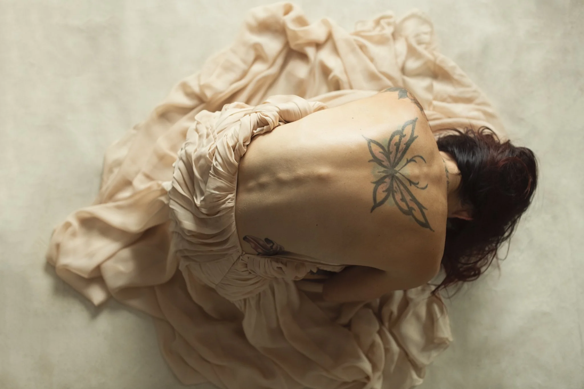 A woman sitting on the floor with her back facing the camera, wearing a loose, beige dress that is gathered around her waist, revealing her back and tattoos including a large butterfly on her upper back and a smaller design on her side.