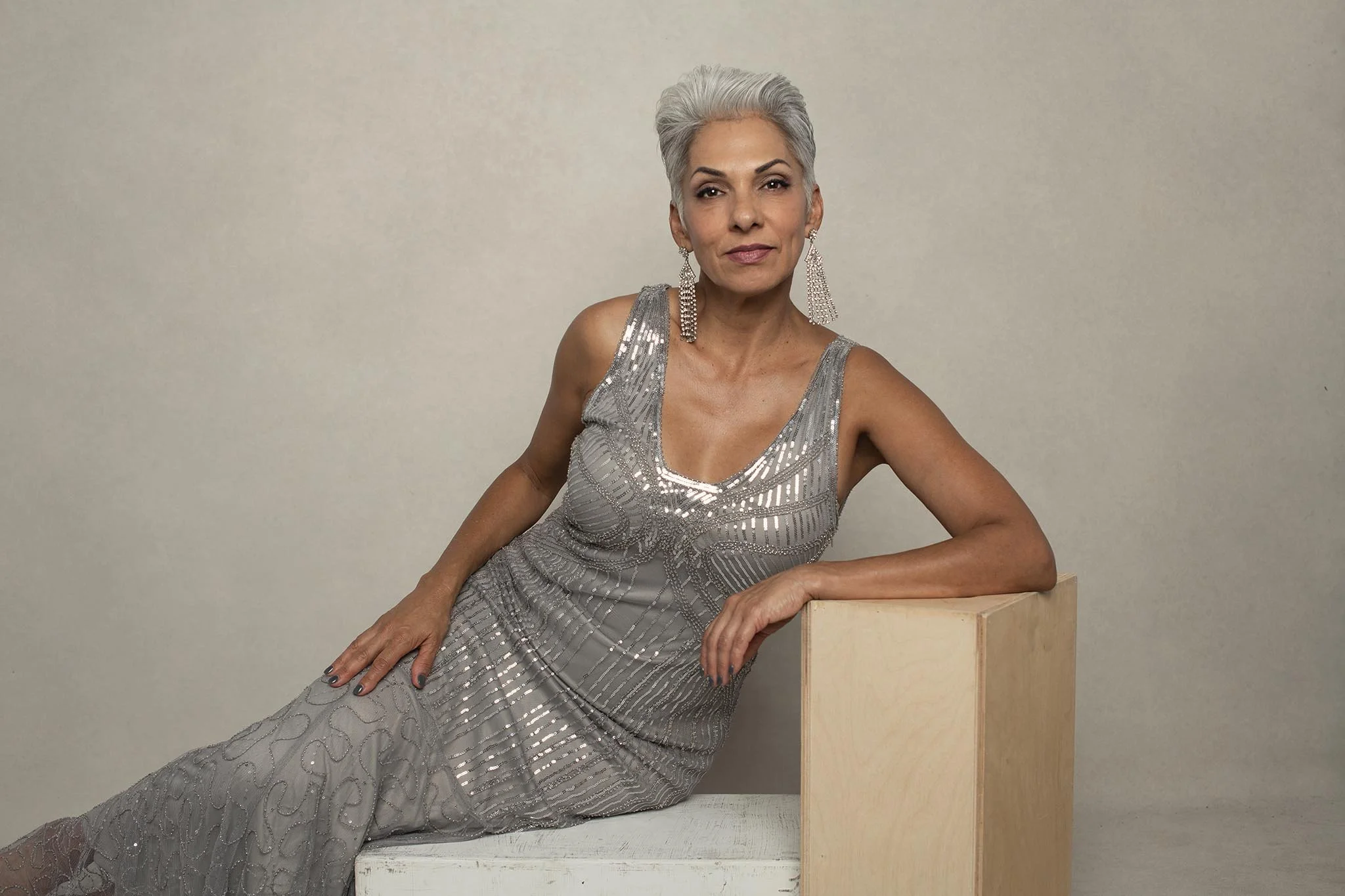 An elegant older woman with silver hair sitting on a white bench, wearing a silver sequined dress and large dangling earrings.