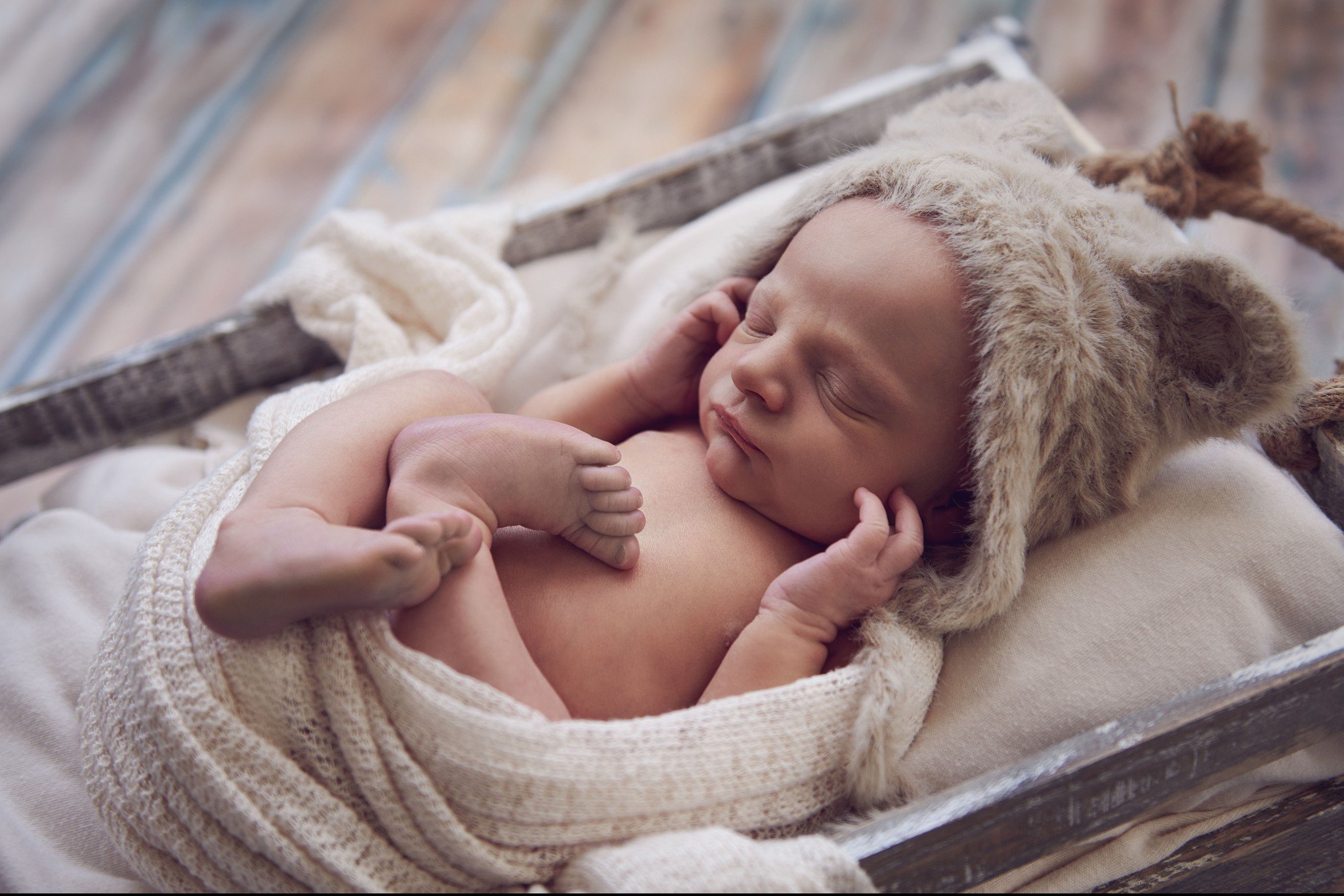 A sleeping baby wearing a fluffy hat with animal ears, lying in a rustic wooden cradle with a soft blanket.
