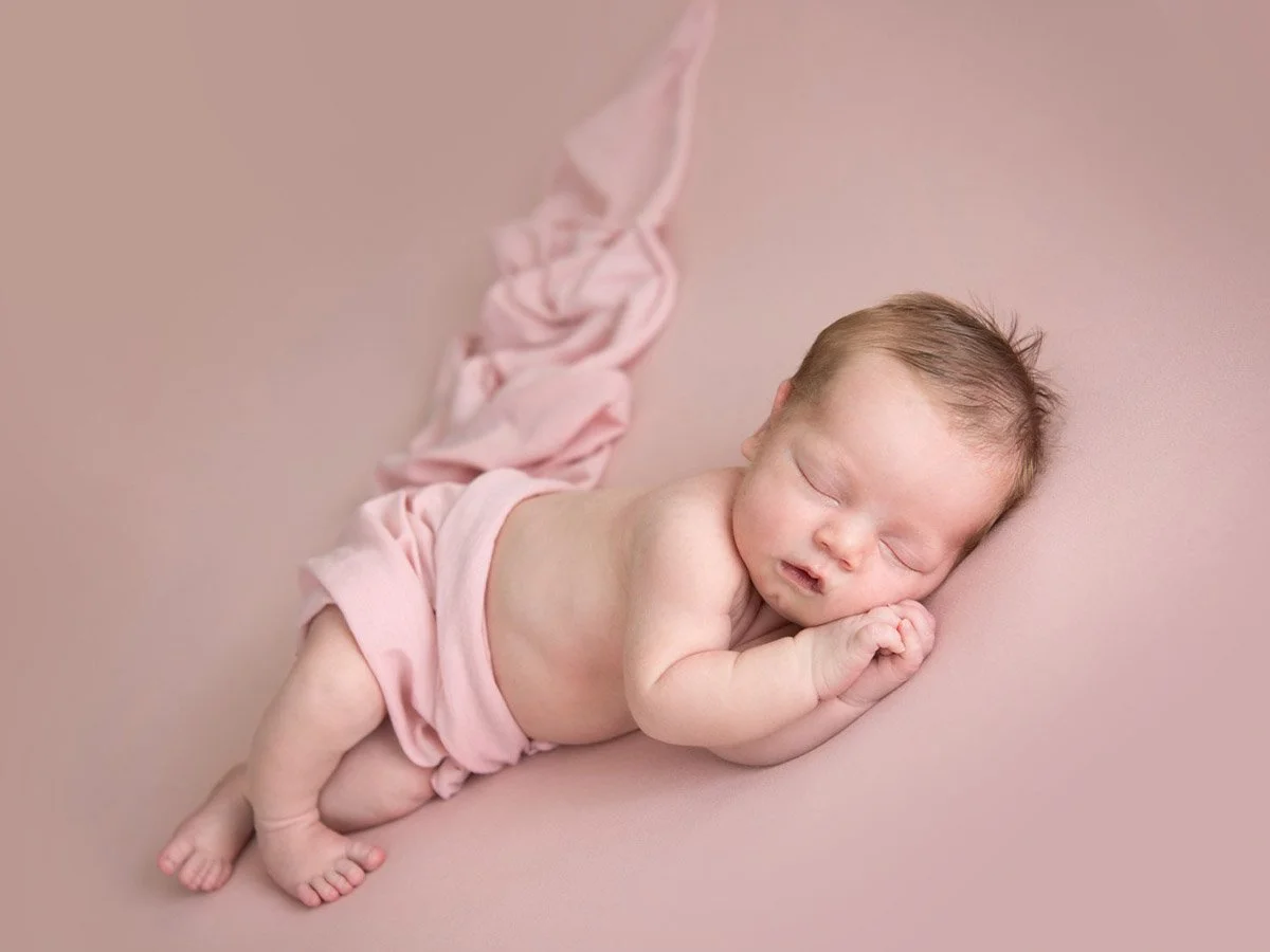 A sleeping newborn baby lying on its side on a pink surface, partially covered with pink cloth.