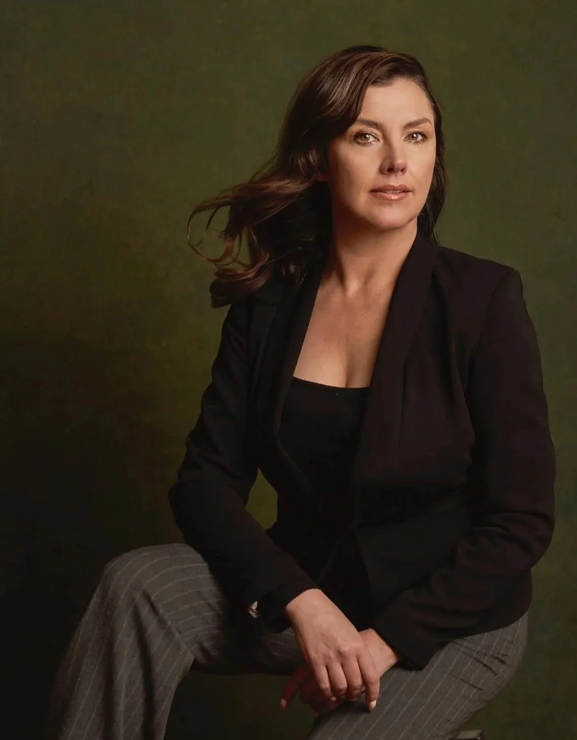 A woman with brown hair, wearing a black blazer and gray striped pants, sitting against a dark green background.