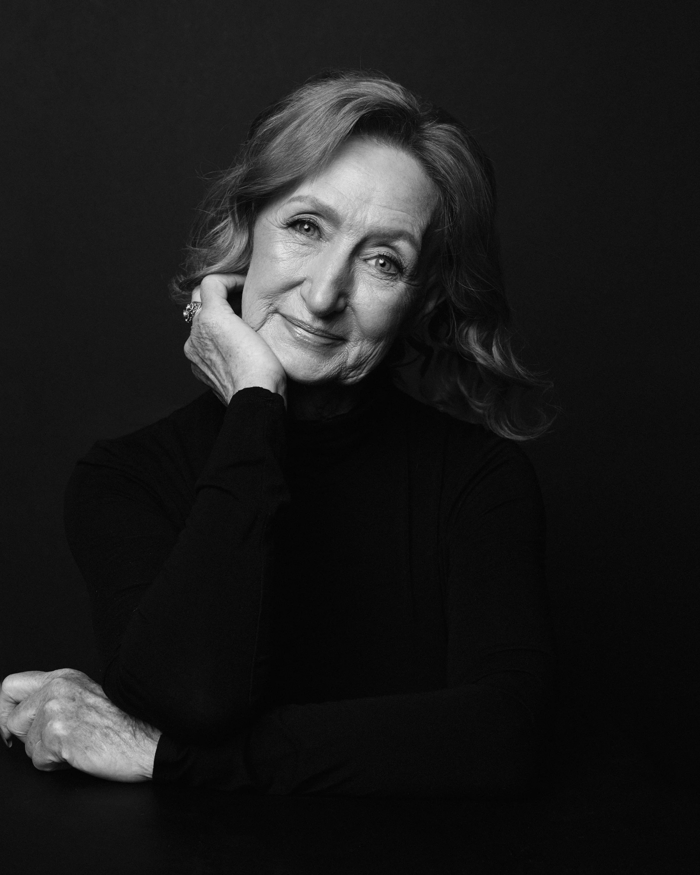 Black and white close-up portrait of an elderly woman with wavy hair, smiling softly, with her hand resting on her face, against a dark background.