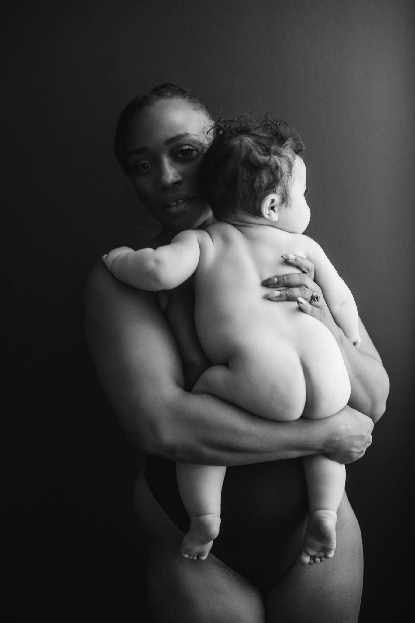 A woman holding a naked child with curly hair, against a dark background.