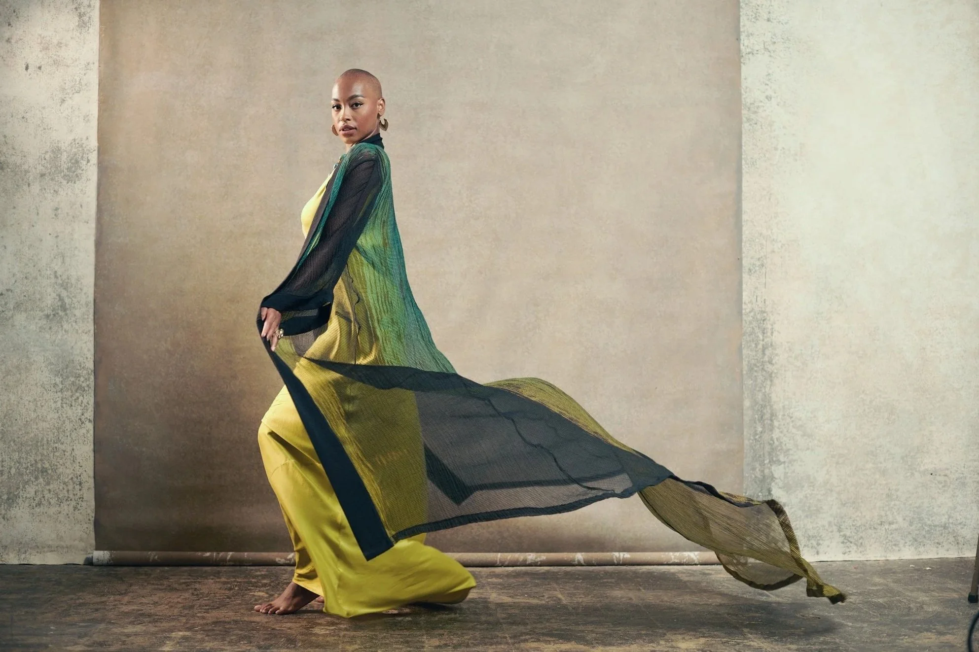 A woman with a shaved head wearing a yellow dress, layered with a sheer, green and black flowy cape, standing against a plain beige wall with a textured appearance.