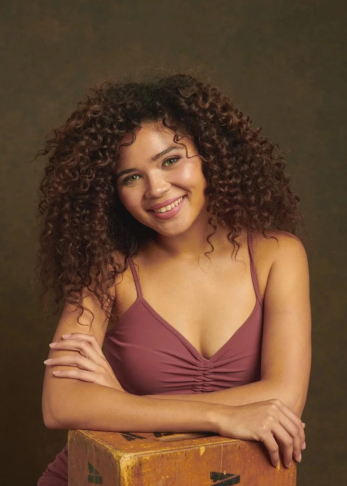 Young woman with curly hair smiling and sitting with arms crossed on a vintage wooden box against a dark background.