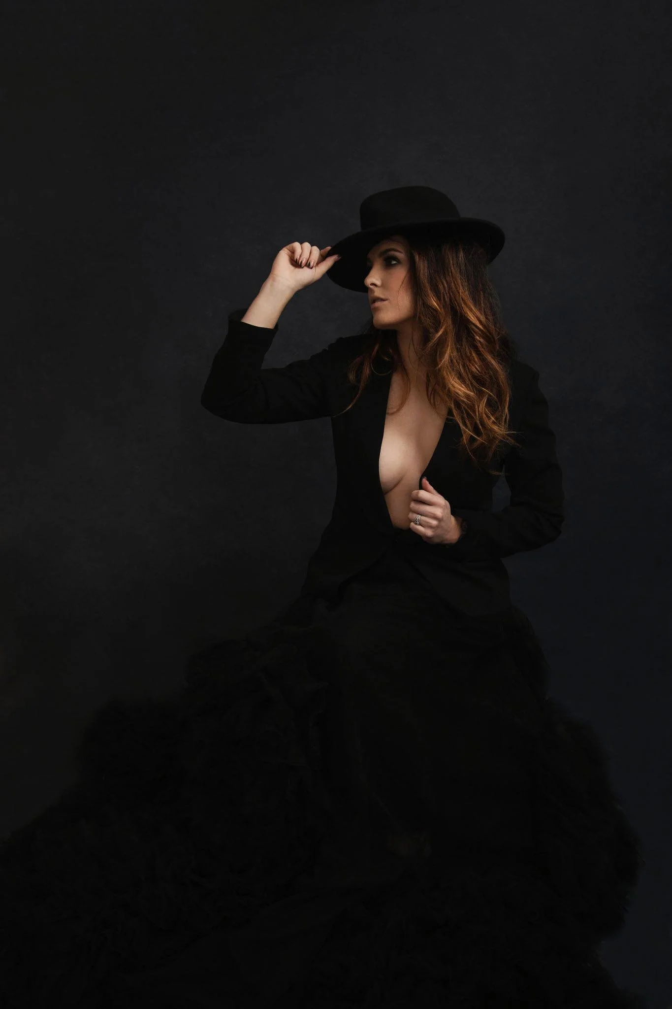 Woman with long red hair, wearing a black hat and black outfit, posing against a dark background.