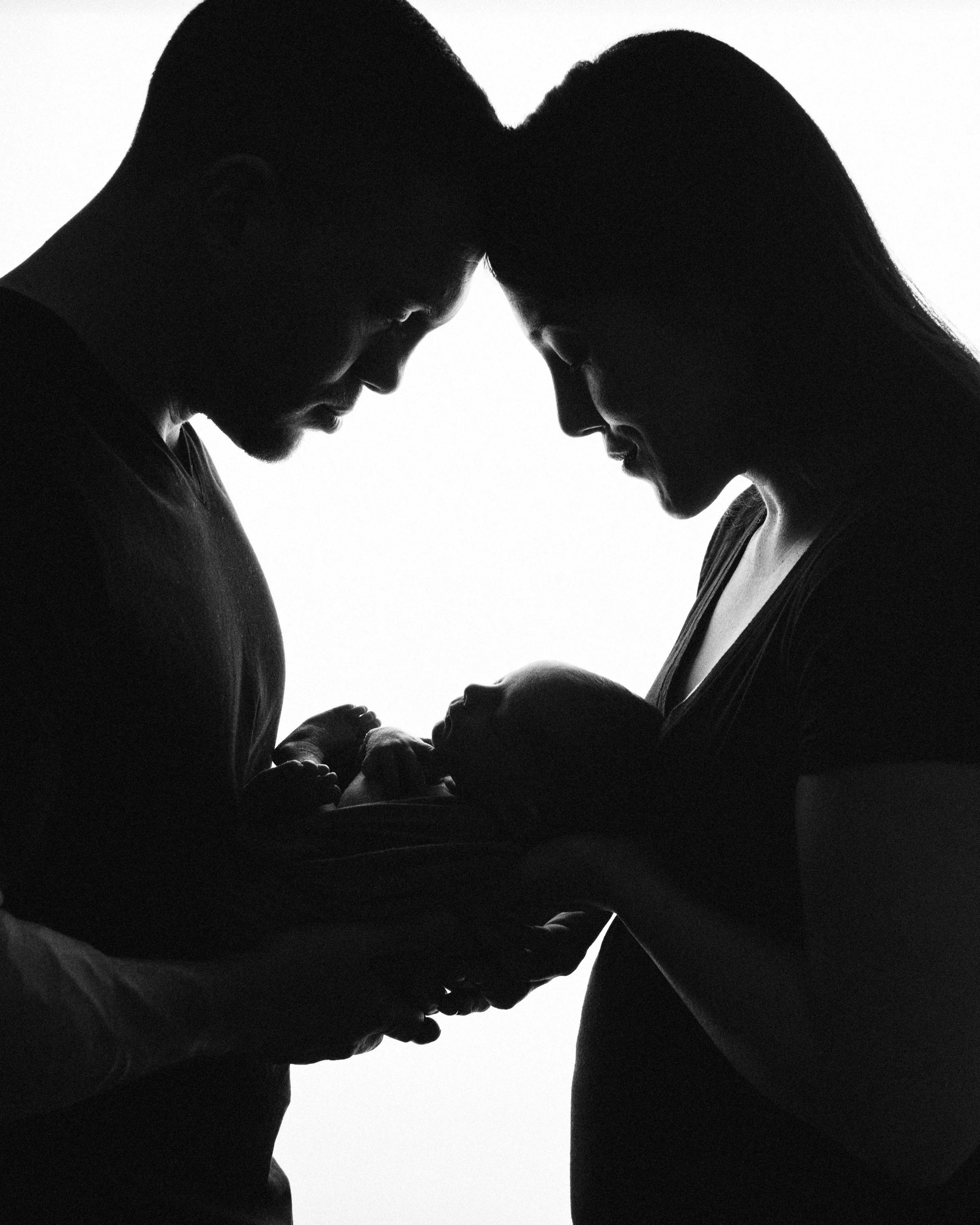 Silhouettes of a man and woman holding a newborn baby, their foreheads touching, in a tender family moment.