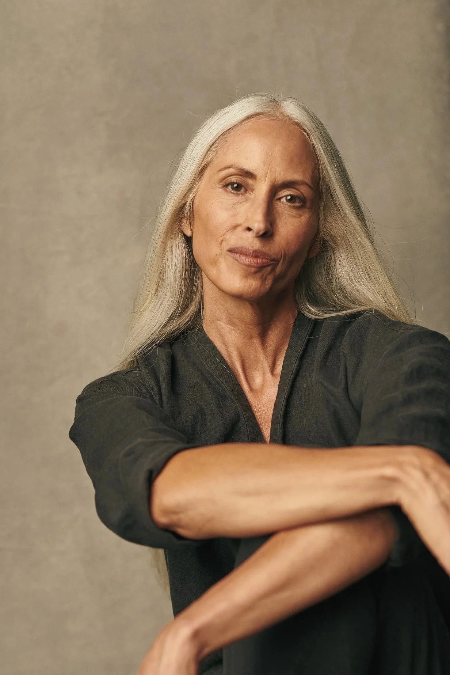 An older woman with long gray hair, wearing a black top, sitting against a neutral background, looking at the camera with a subtle smile.