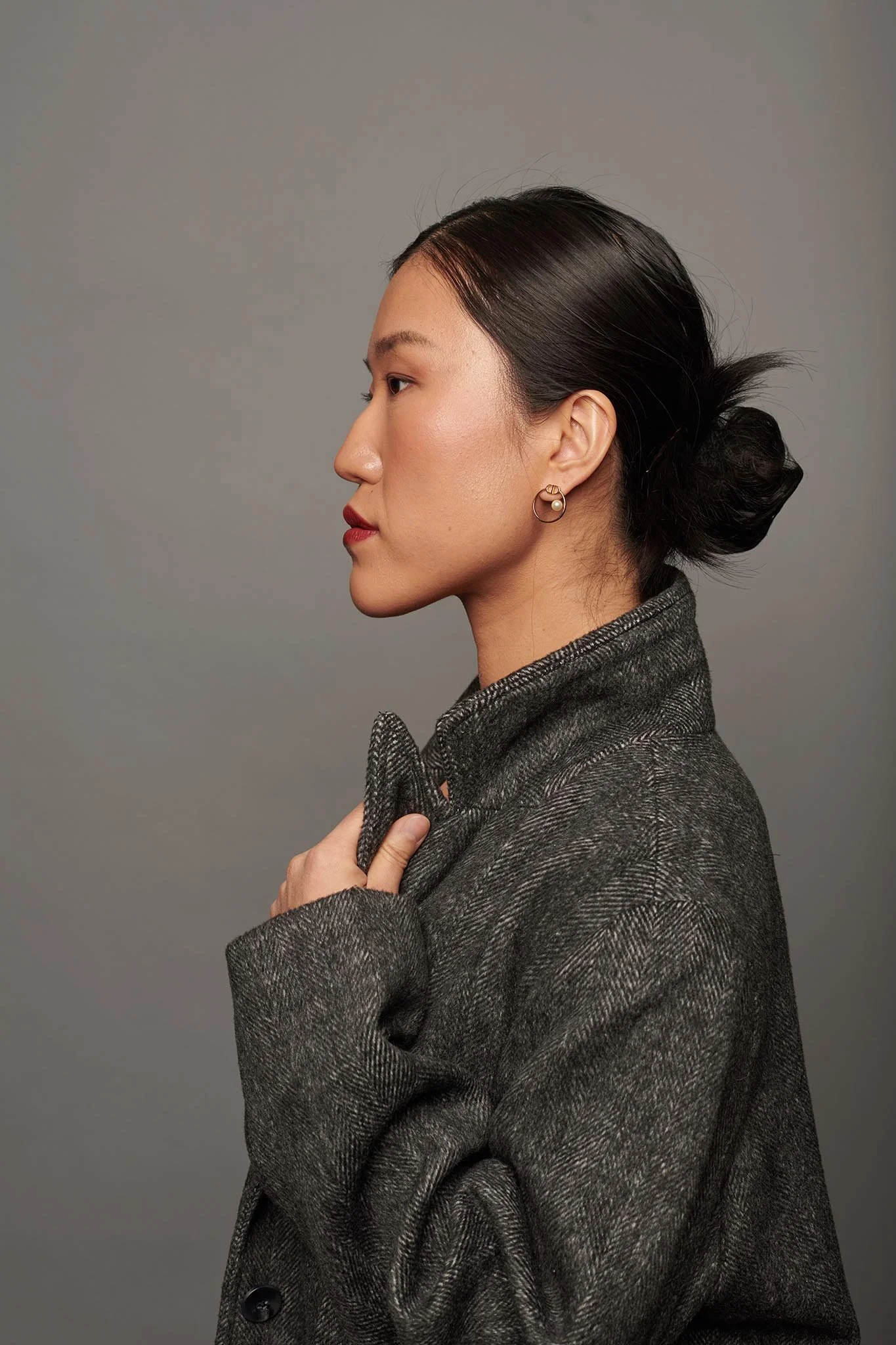 A side profile of a woman with dark hair in a bun, wearing a grey textured coat and small hoop earrings against a grey background.