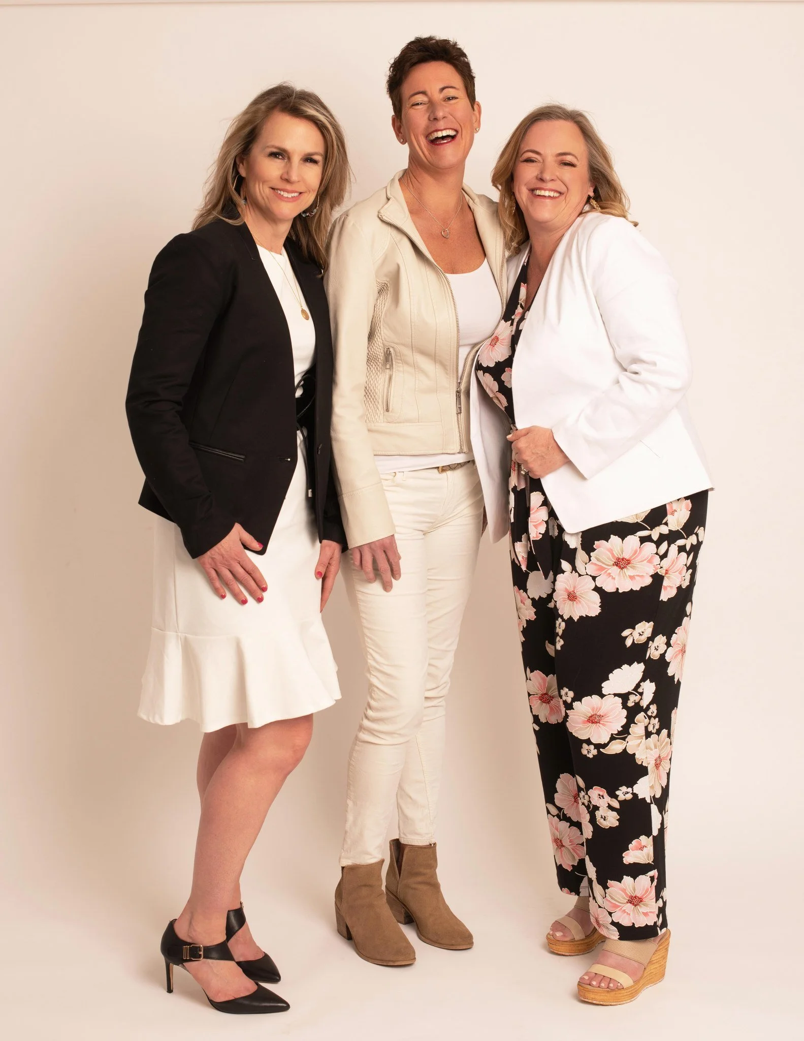 Three women standing together, smiling, against a plain background. All are dressed in stylish clothing and appear happy.