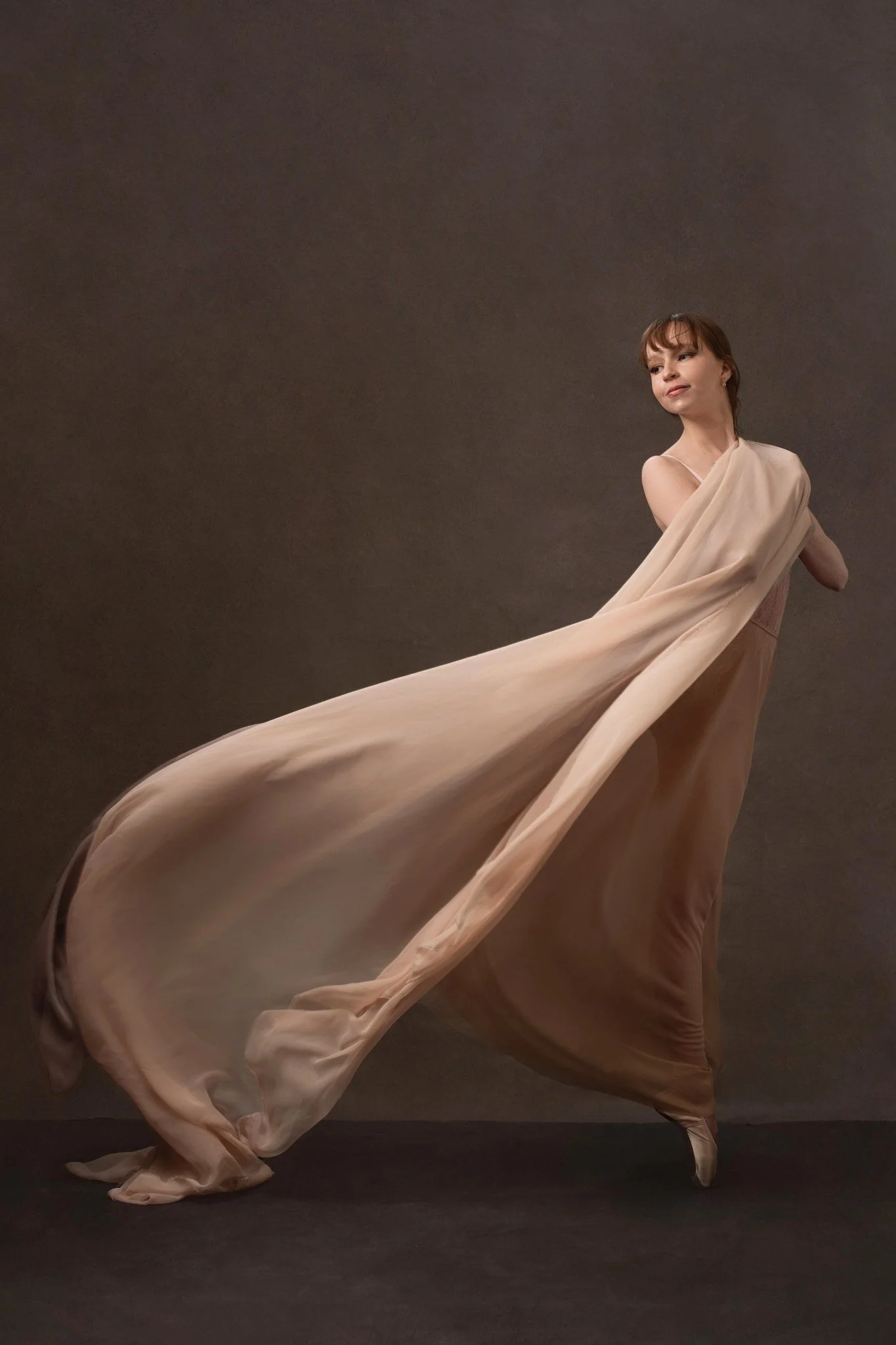 A young woman in a beige gown with an attached flowing train posing against a plain background.