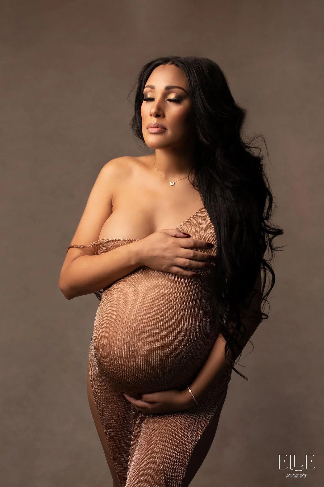 A pregnant woman with long black hair and makeup, wearing a sheer, skin-colored dress that reveals her baby bump, posing with her eyes closed and hand on her chest against a neutral background.