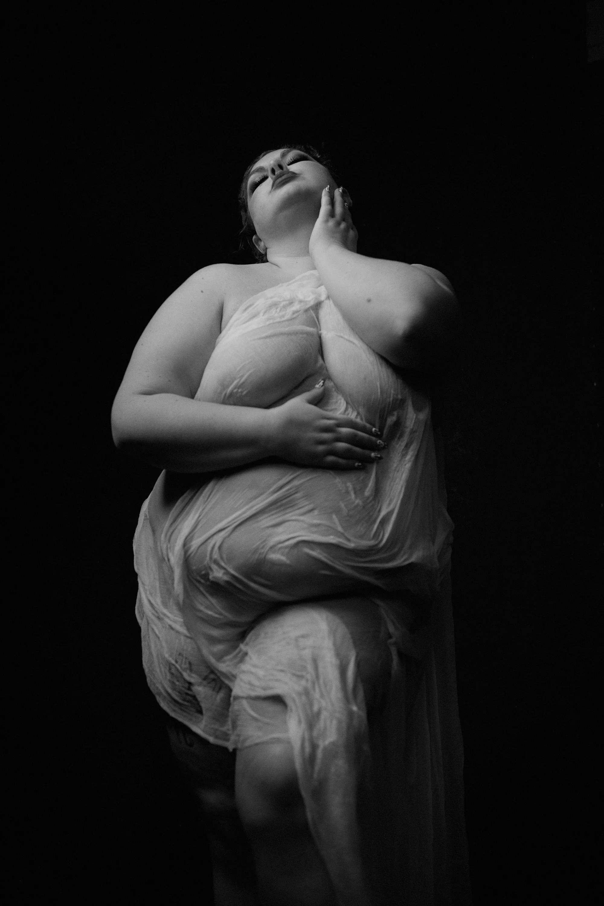 Black and white portrait of a woman in a flowing dress with her eyes closed, head tilted back, one hand on her chin and the other on her stomach, against a dark background.