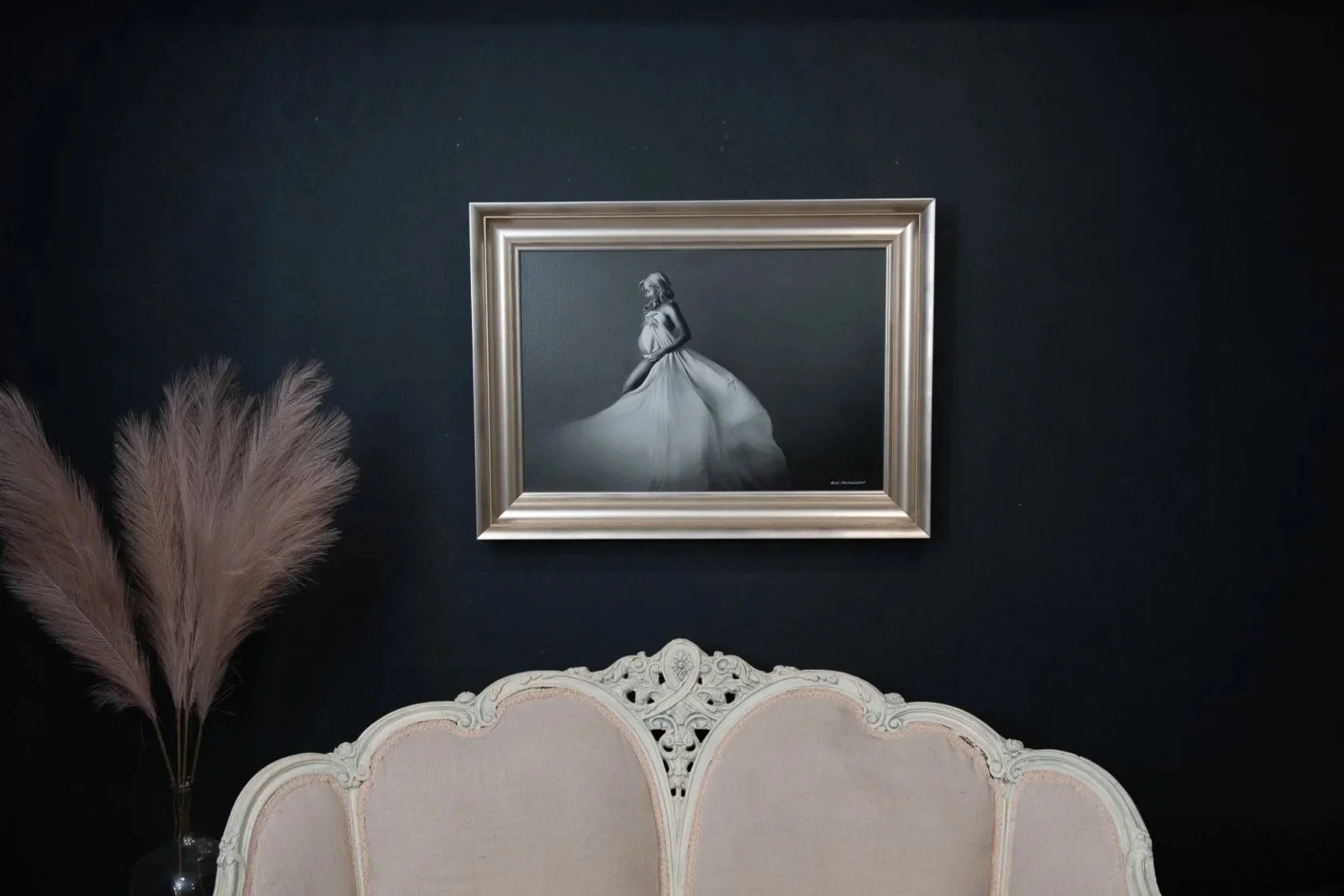 A painting of a woman in a flowing dress hanging on a dark wall above a beige upholstered headboard, with pampas grass in a vase to the left.