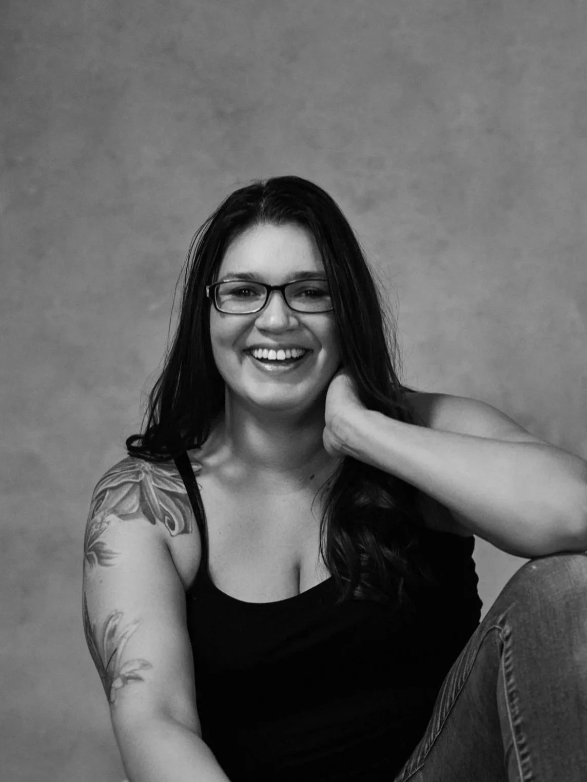 Black and white photo of a smiling young woman with glasses, long dark hair, a tattoo on her left arm, wearing a sleeveless top, sitting with one arm resting behind her neck against a plain background.