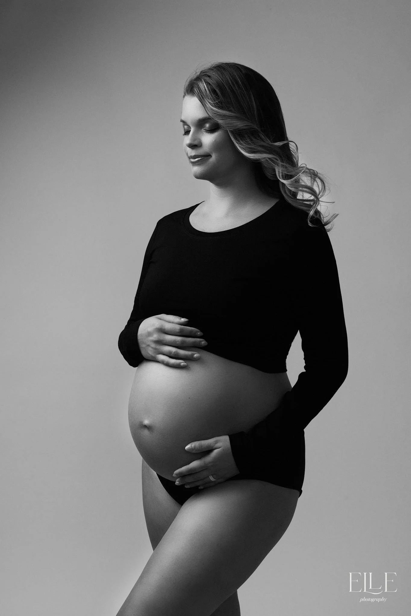 Black and white photo of a pregnant woman with closed eyes, smiling slightly, with her hands gently resting on her belly, wearing a long sleeve black top and underwear, against a plain background.