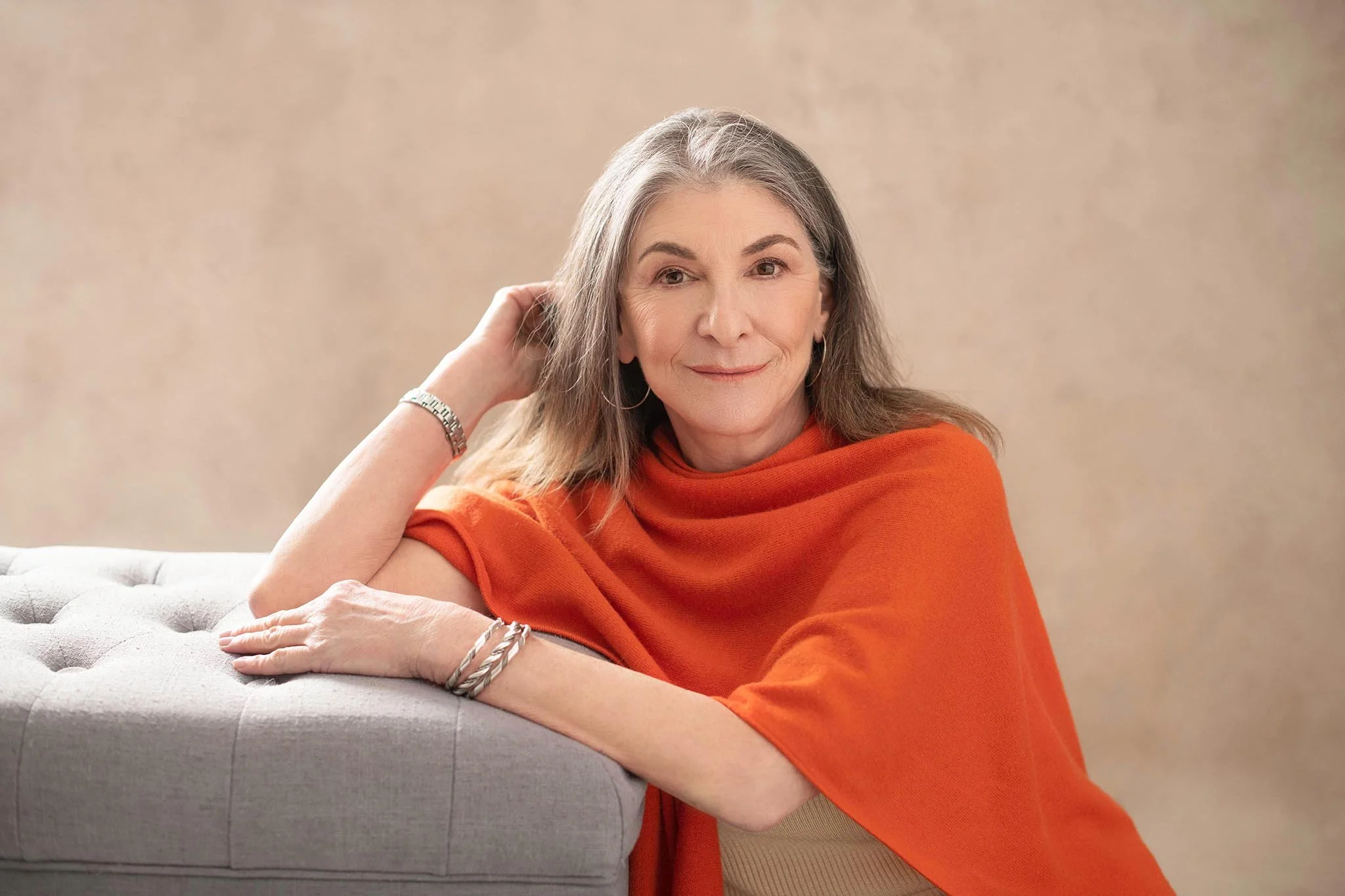 A smiling elderly woman with gray hair and earrings, wearing an orange top, resting her chin on her hand, sitting on a light-colored tufted couch against a neutral background.