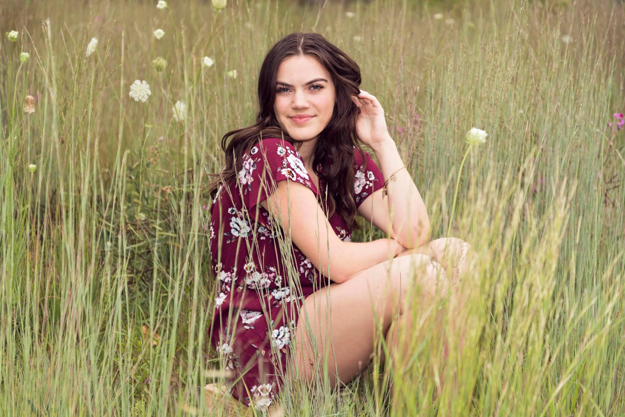 A young woman with long brown hair and blue eyes sitting in a grassy field, wearing a red floral dress, smiling softly, with her hand touching her hair.