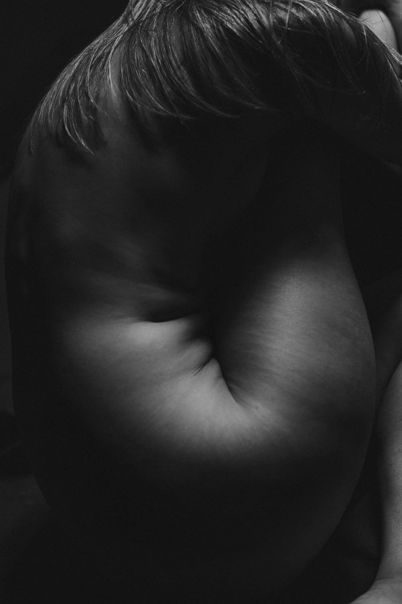 Black and white photograph of a person with their head down, revealing their back and shoulder, with short hair visible.