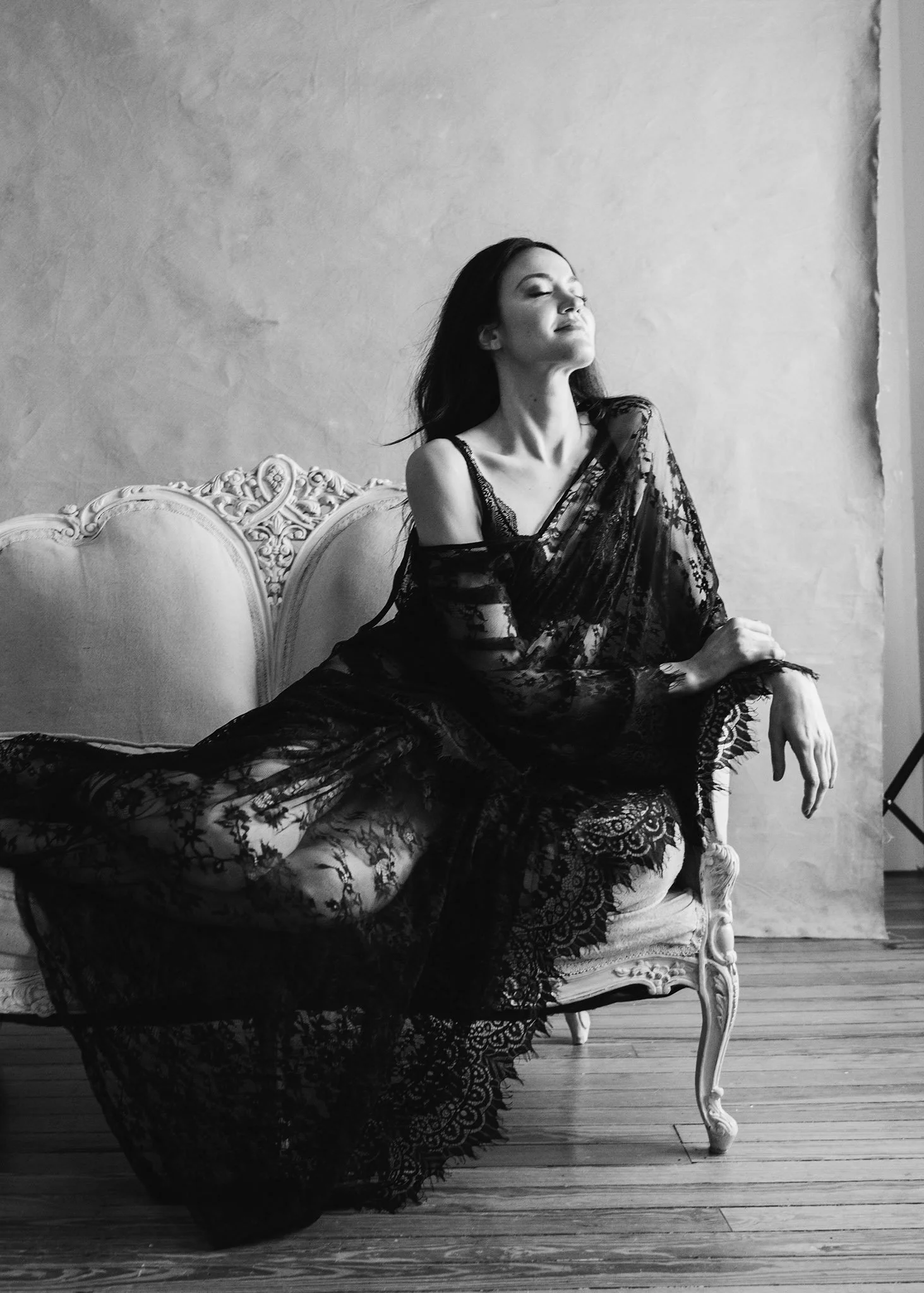 A woman sitting on an ornate vintage couch, wearing a lace black dress, with her eyes closed and a serene expression, in a room with a plain wall background.