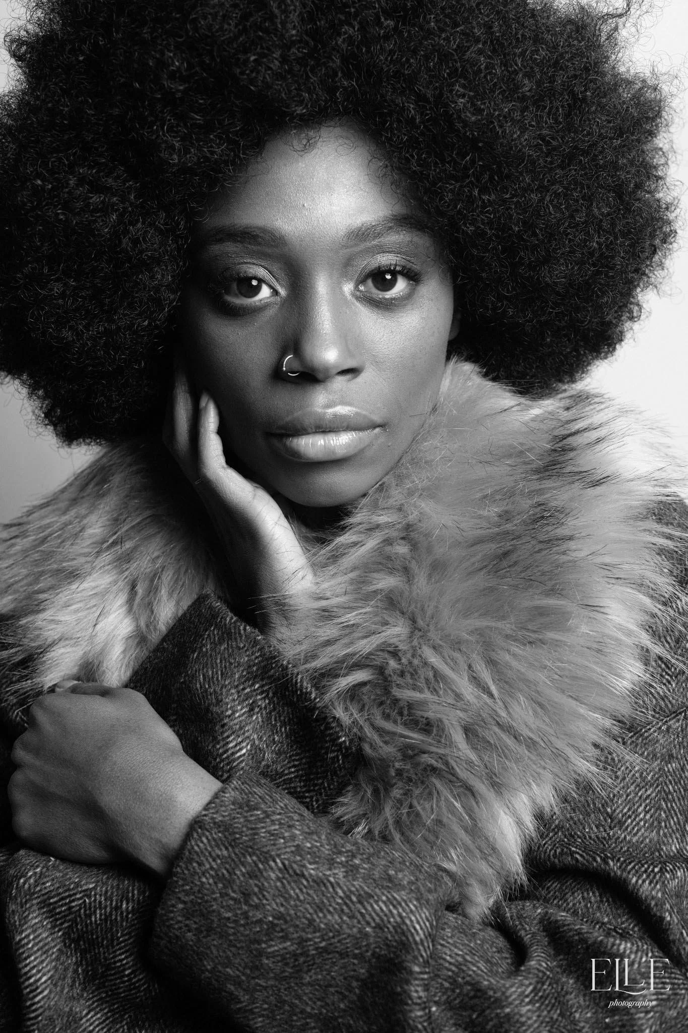 A black and white portrait of a woman with a large, curly afro hairstyle, wearing a nose ring, a fur collar, and a textured coat, looking directly at the camera with her hand gently touching her face.