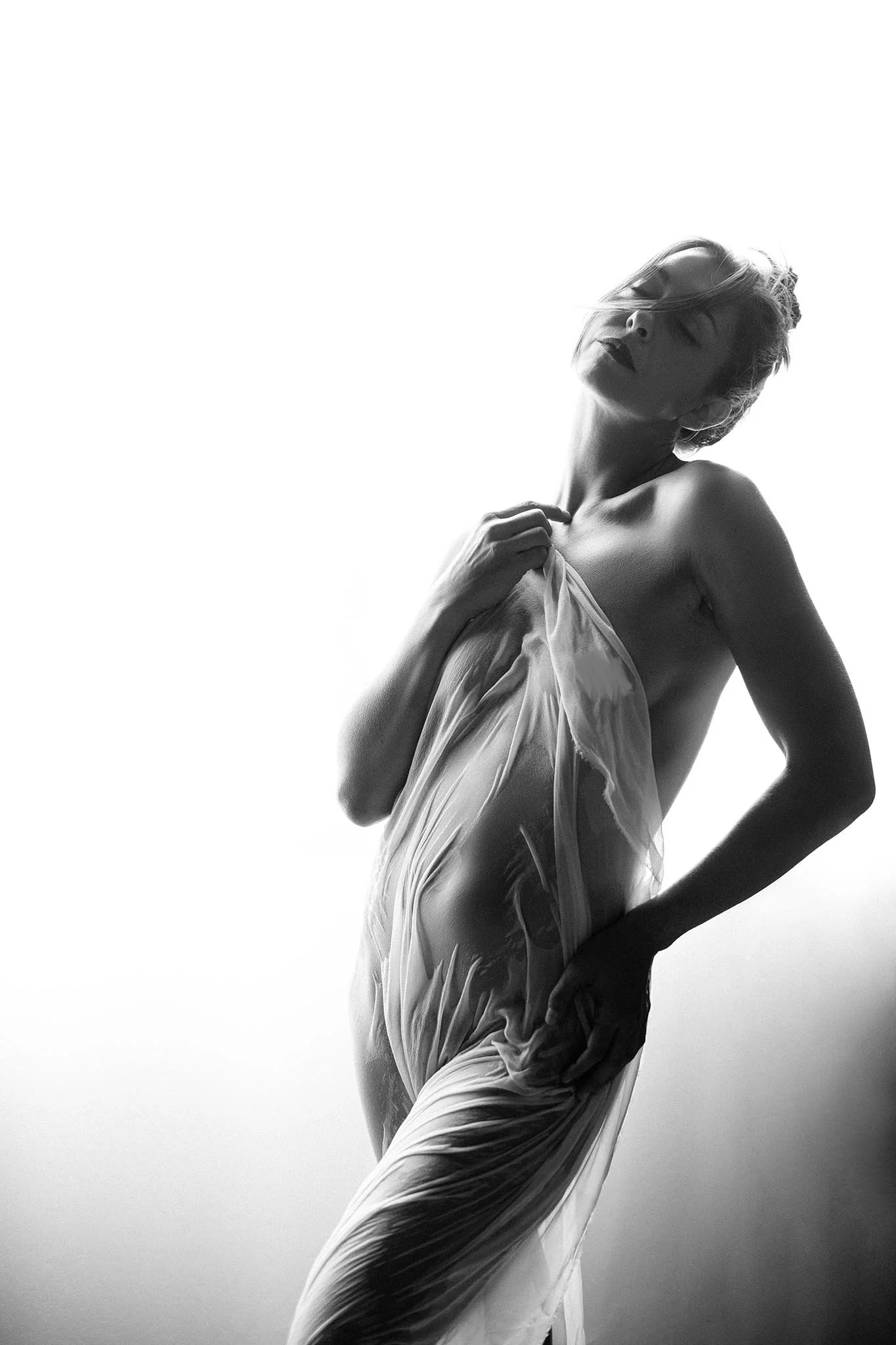 Black and white photo of a woman with her eyes closed, holding a sheer fabric against her chest with one hand and placing the other on her hip.