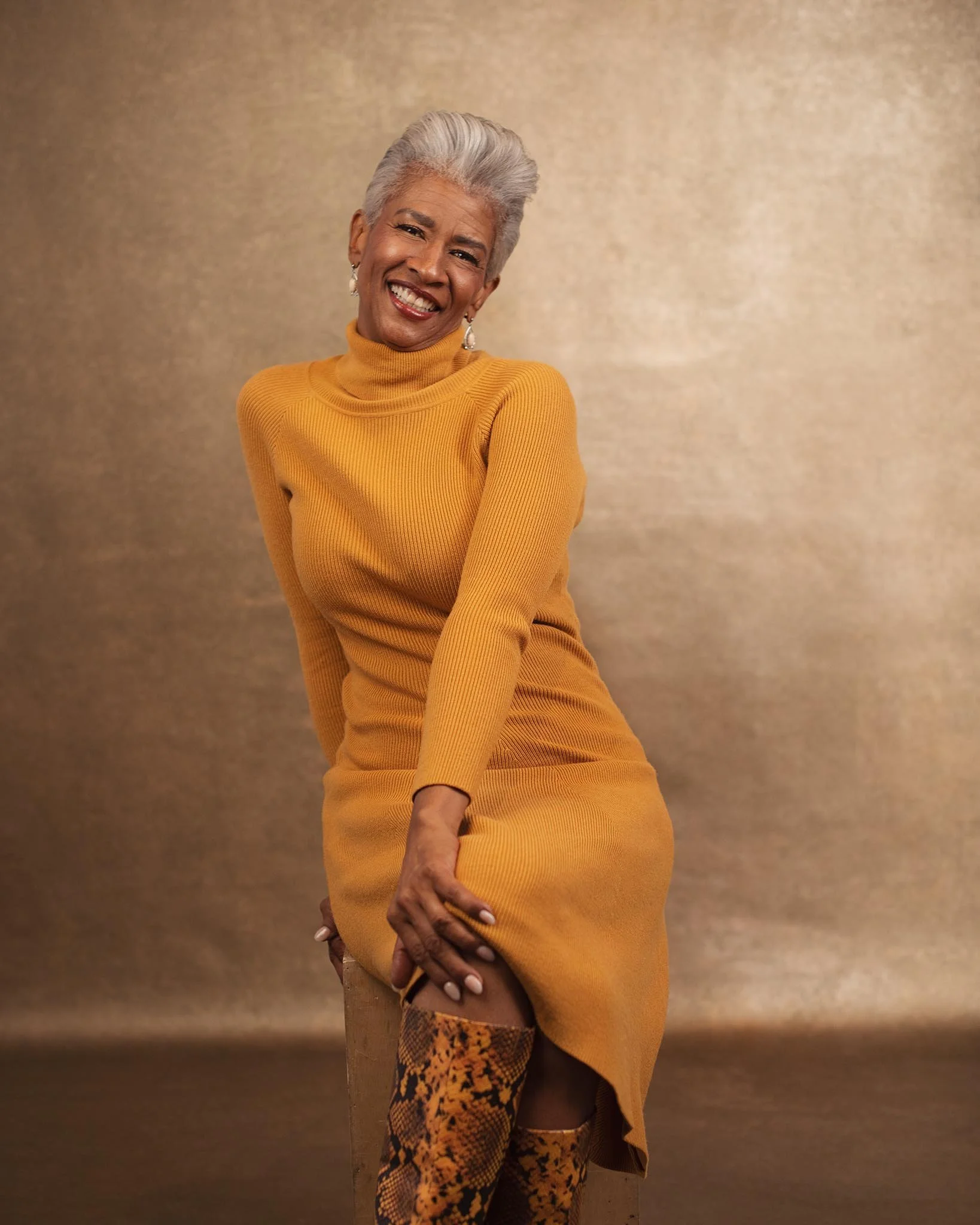 A smiling woman with short gray hair, wearing a mustard-colored turtleneck sweater dress and snakeskin-patterned thigh-high boots, sitting against a gold background.
