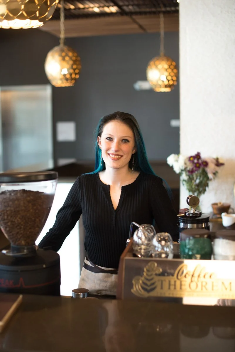 A smiling woman with dark and teal hair behind a coffee bar with coffee equipment, flowers, and coffee beans in a coffee shop.