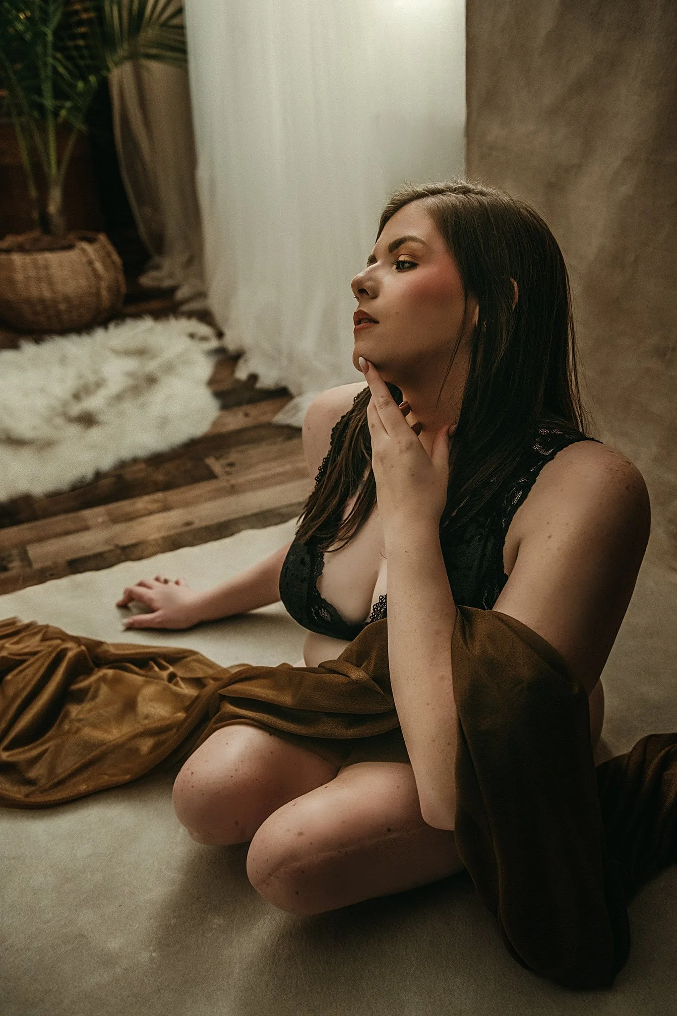 A woman with dark hair styling sitting on the floor in a photo studio, wearing black lace lingerie and a brown shirt, with a contemplative pose, in a warmly lit room with a throw rug and plant in the background.