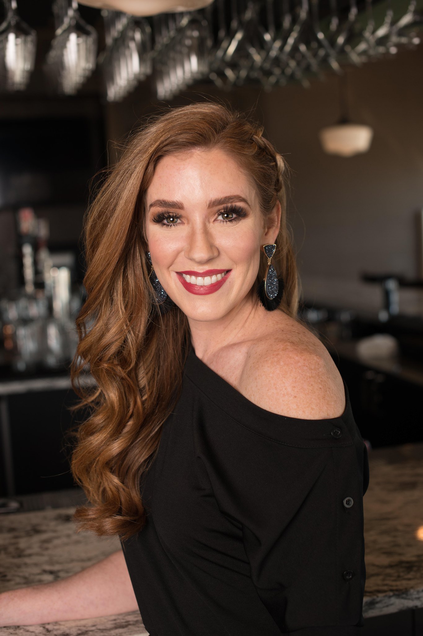 A smiling woman with red hair and makeup, wearing large earrings and an off-the-shoulder black top, in a bar or restaurant setting.