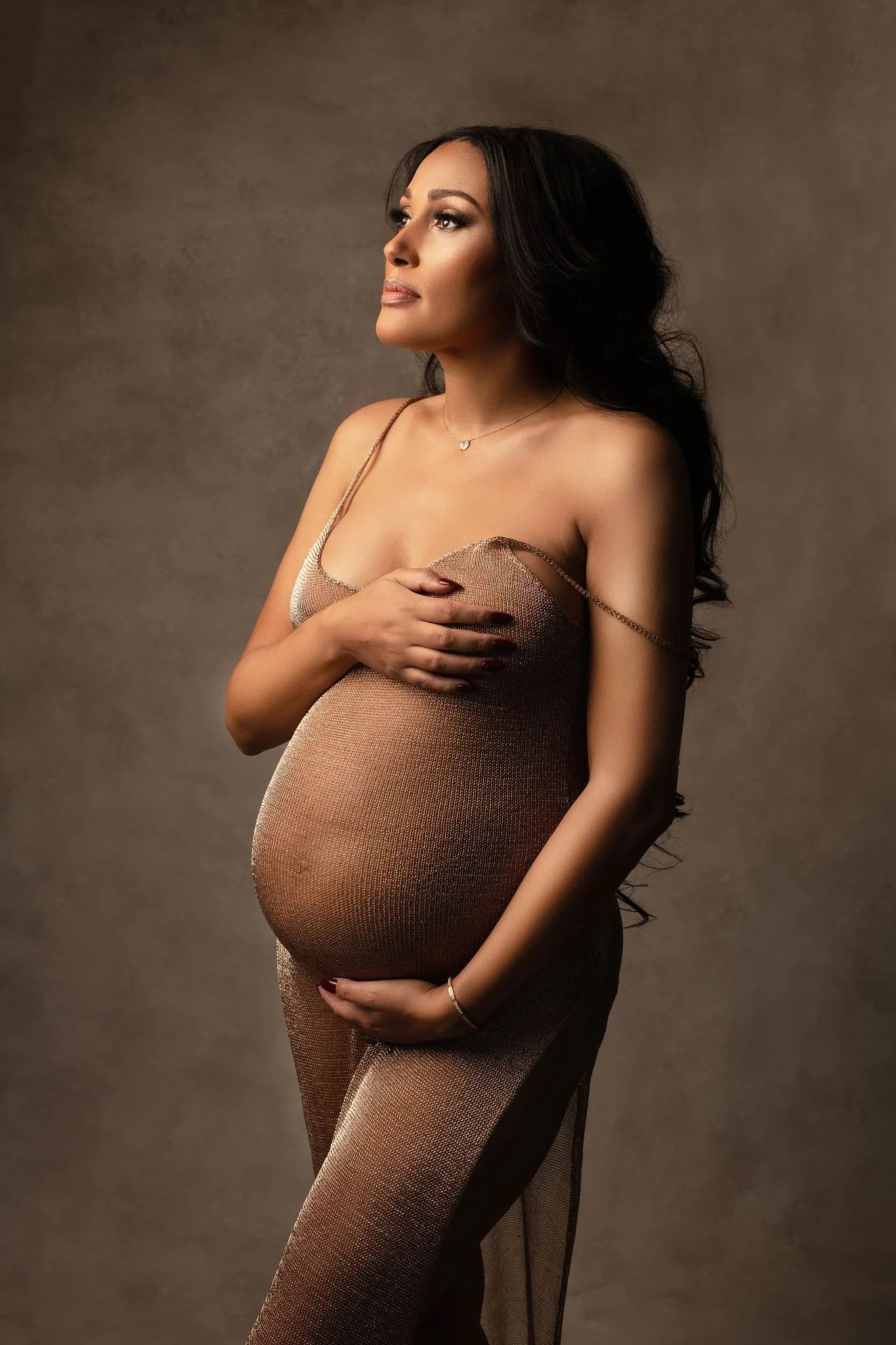 Pregnant woman with long dark hair in gold dress, holding her belly with one hand and her chest with the other, facing side profile against a neutral background.