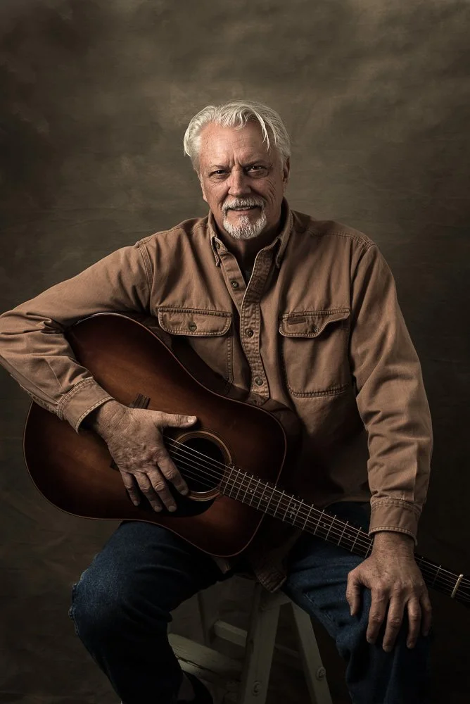 Older man with white hair and beard sitting on a stool, holding an acoustic guitar, wearing a tan button-up shirt and blue jeans, against a dark studio background.