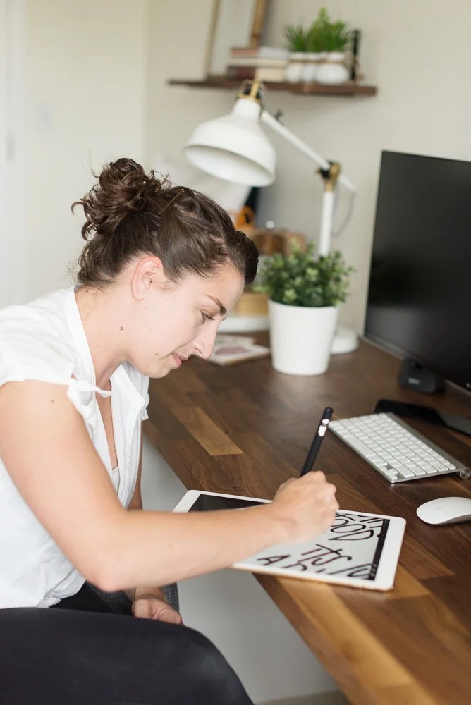 Woman drawing black and white graphics on a tablet with a stylus at a wooden desk, surrounded by a computer, keyboard, mouse, and potted plants.